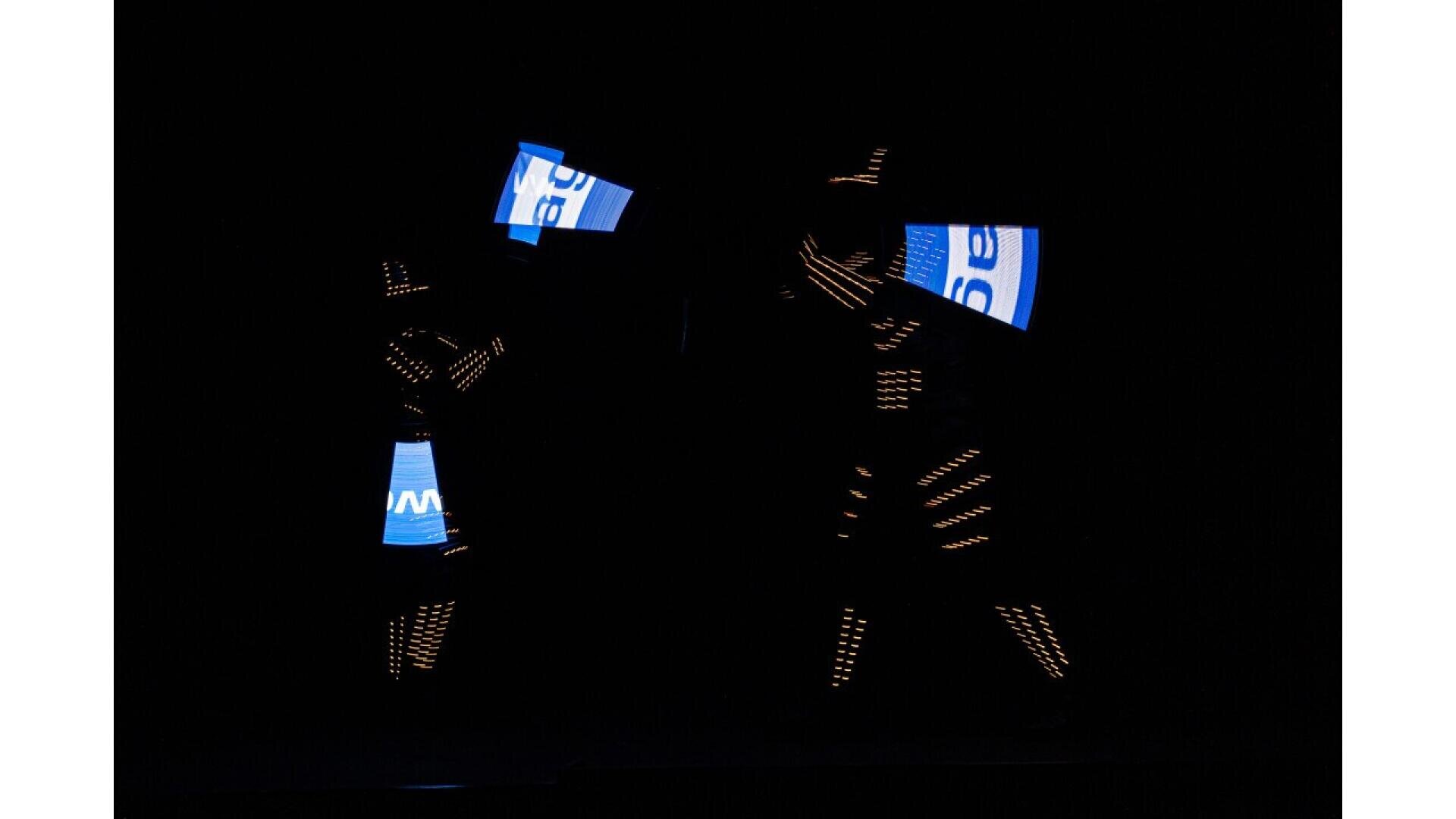 Two performers in dark suits with glowing LED lights hold bright blue and white rectangular panels, standing against a black background, creating a striking visual effect.