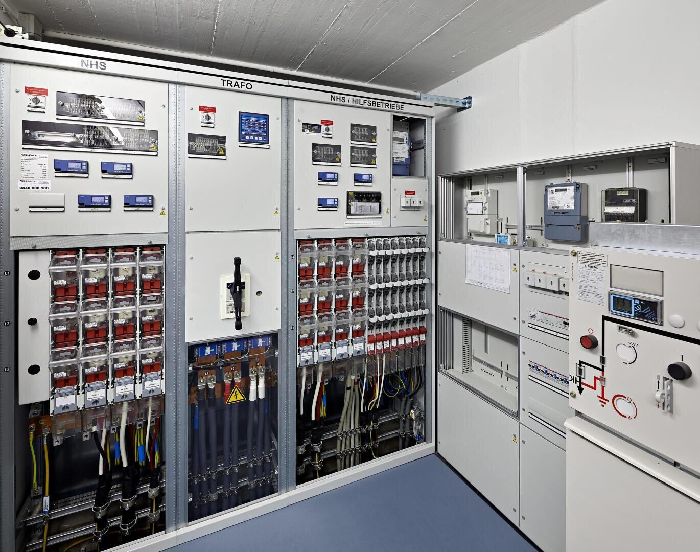 Elektrischer Kontrollraum mit mehreren Schalttafeln, Stromkreisen, Messgeräten und Schaltern an den Wänden. Der Raum ist sauber, gut beleuchtet und organisiert, mit beschrifteten Geräten und sichtbaren Kabeln unter den Schalttafeln.