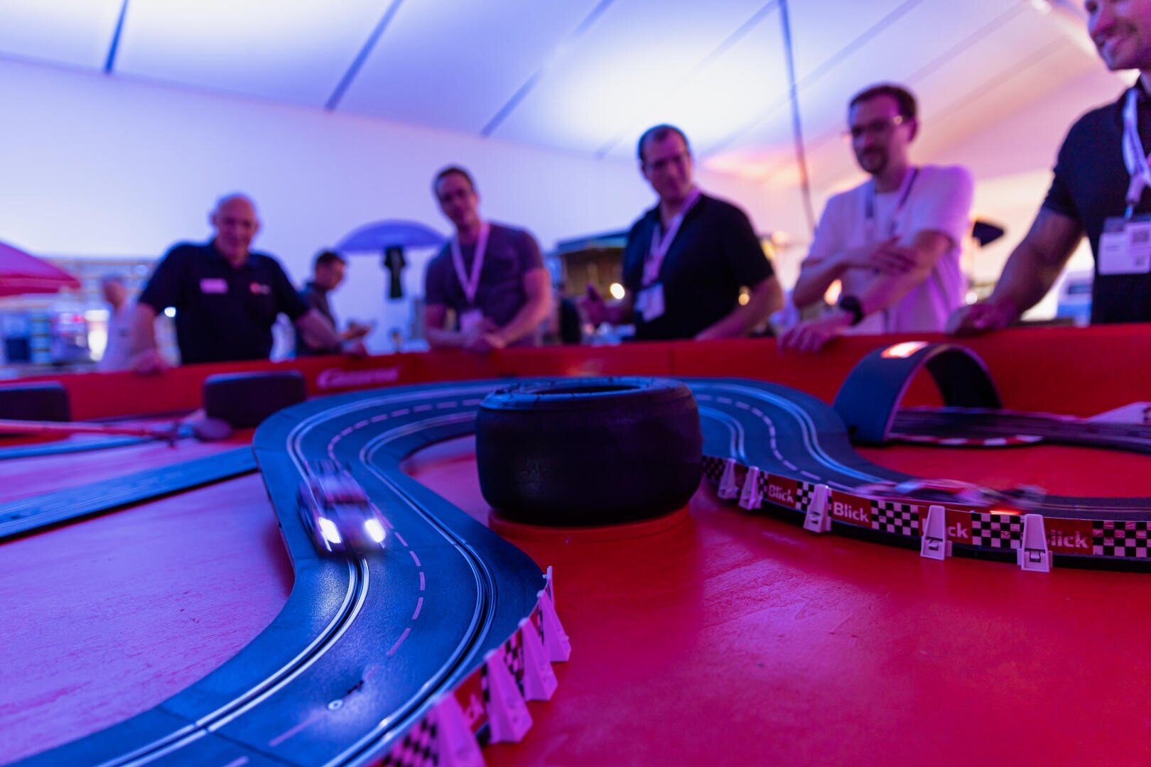Eine Gruppe von Menschen lächelt und beobachtet, wie Spielzeugautos auf einer kurvenreichen Strecke bei einer Indoor-Veranstaltung um die Wette fahren. Die Szene ist hell beleuchtet mit einem roten und blauen Farbschema.