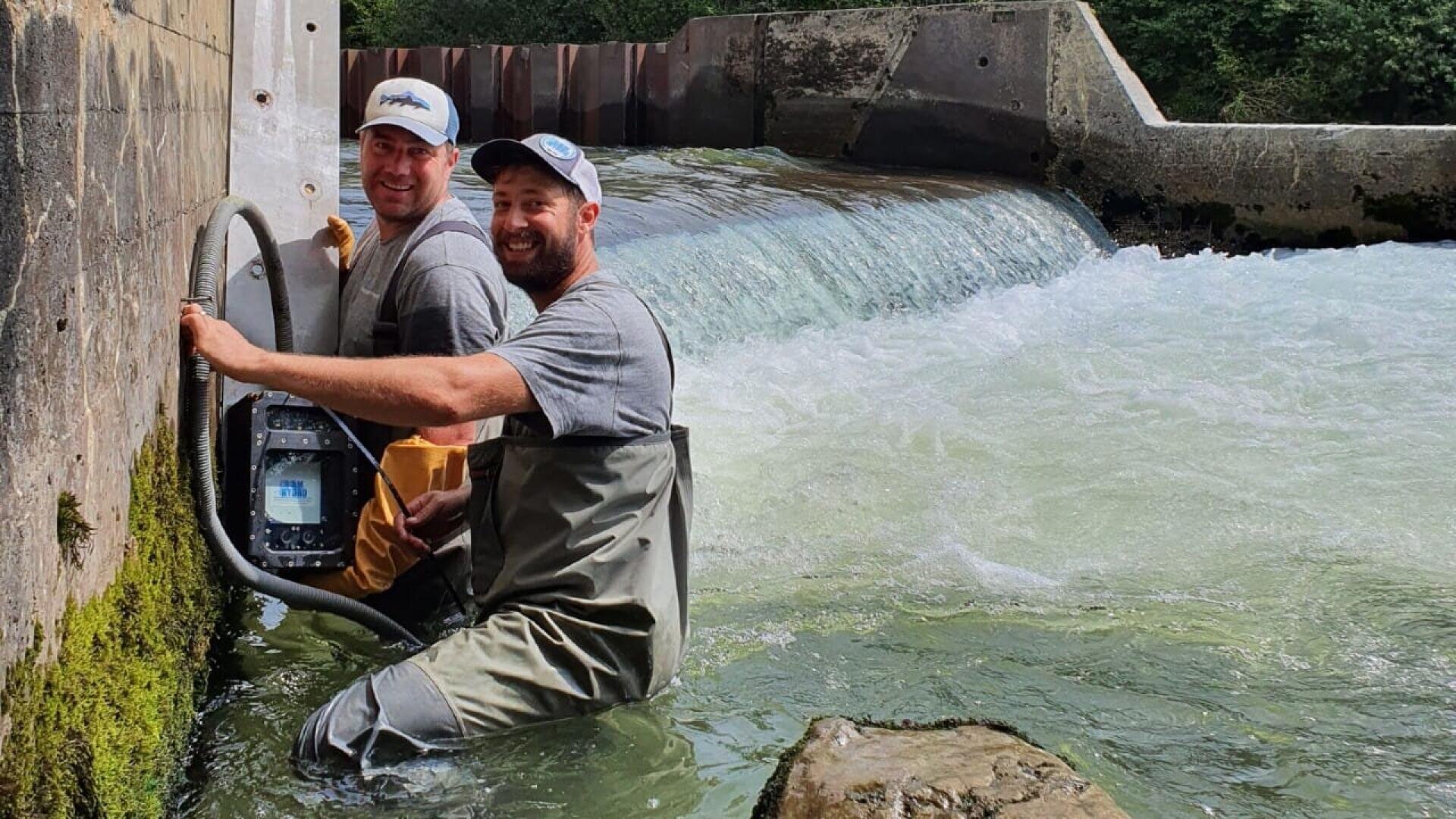 Zwei Männer in Wathosen lächeln, während sie an einer Betonmauer in einem flachen, schnell fließenden Fluss in der Nähe eines kleinen Wasserfalls oder Wehrs Ausrüstung installieren, wobei im Hintergrund Bäume zu sehen sind.