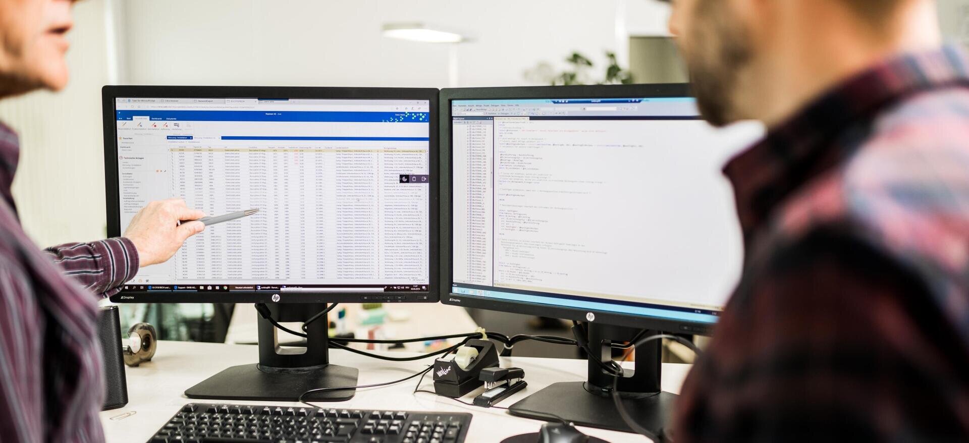 Zwei Personen arbeiten an einem Schreibtisch mit zwei Monitoren; eine Person zeigt auf eine Tabellenkalkulation auf dem linken Bildschirm, während auf dem anderen Bildschirm ein Code angezeigt wird. Beide sind in einer hellen Büroumgebung auf die Computerbildschirme konzentriert.