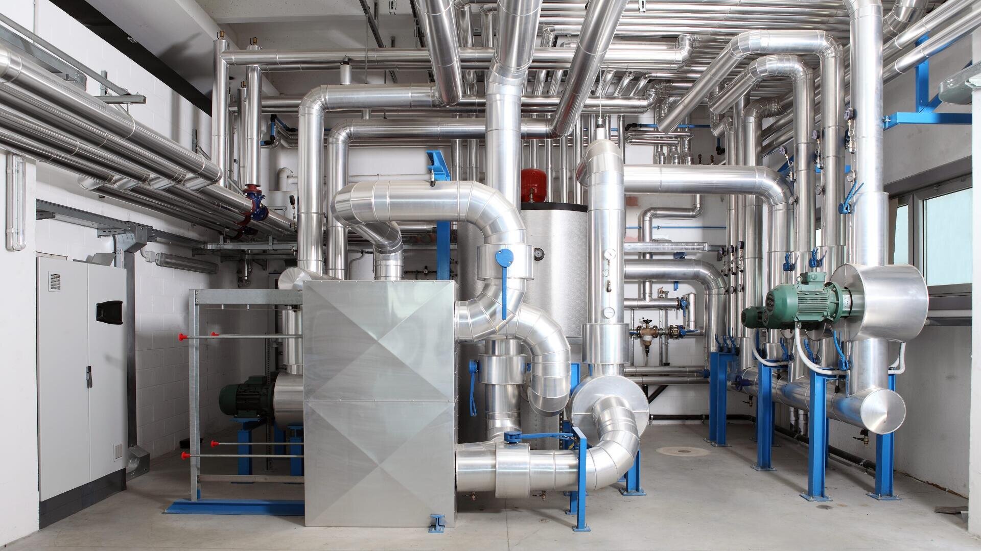 Ein Raum mit Industrieanlagen und Metallrohren, einschließlich großer isolierter Kanäle, Pumpen und Maschinen, die für Heizungs-, Lüftungs- oder Kühlsysteme in einer Gewerbe- oder Industrieanlage verwendet werden.