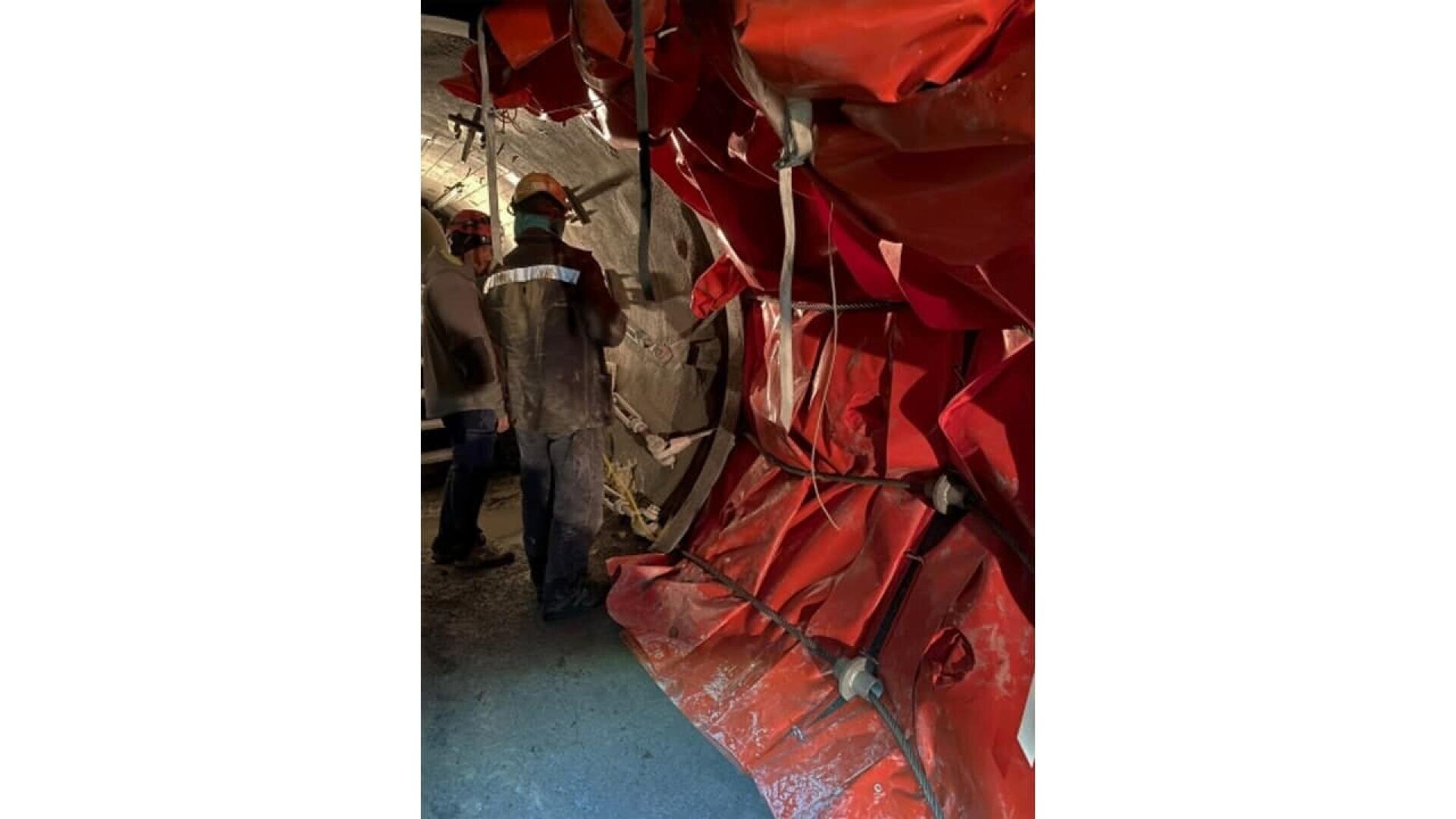 Two workers wearing safety helmets and reflective jackets stand near a large, red, flexible duct or barrier with metal supports inside an industrial or construction setting.