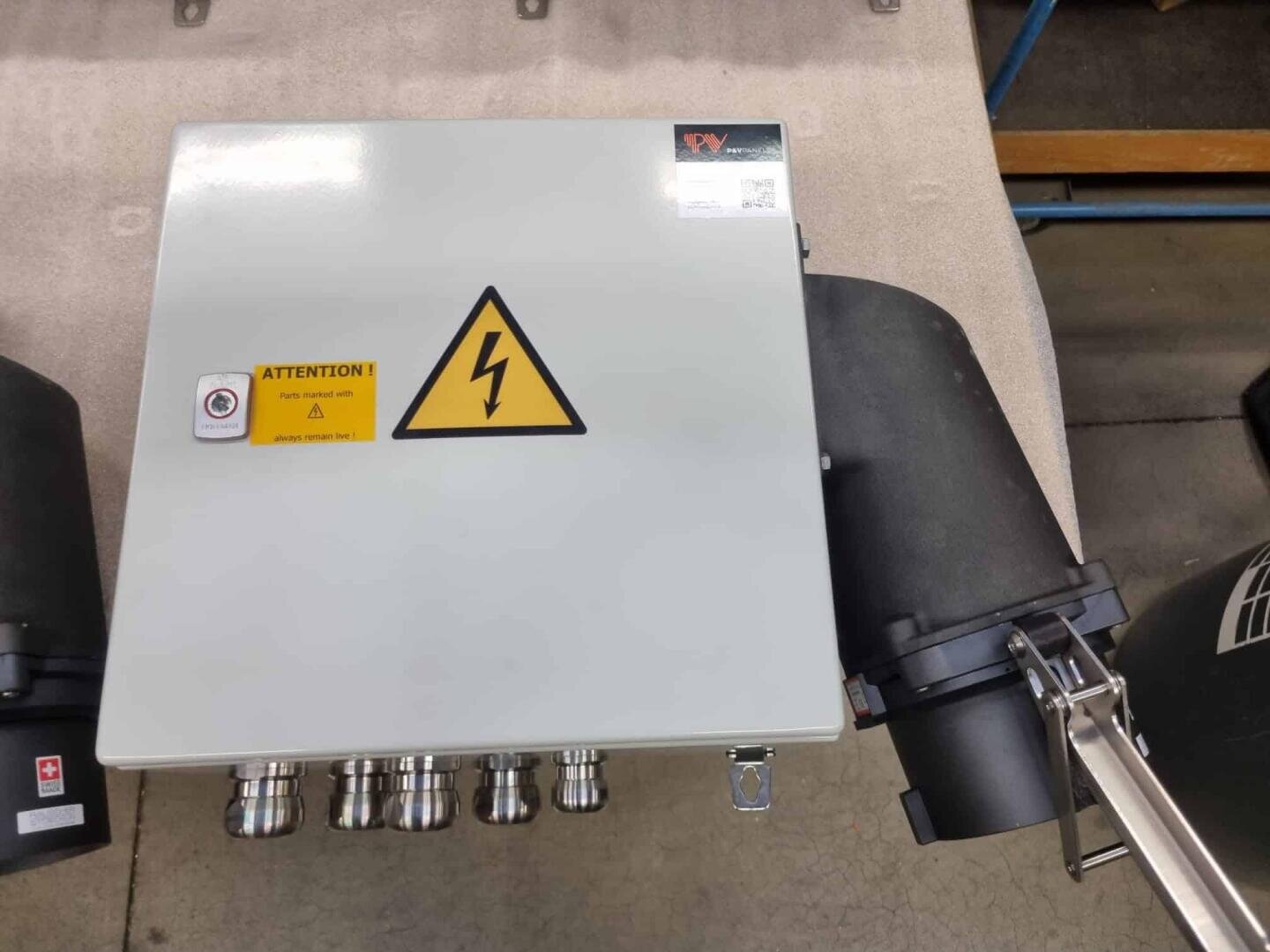 A gray electrical control box with a high voltage warning symbol and an Attention! safety label, mounted on a surface, with multiple cable connectors and black components attached on the right side.