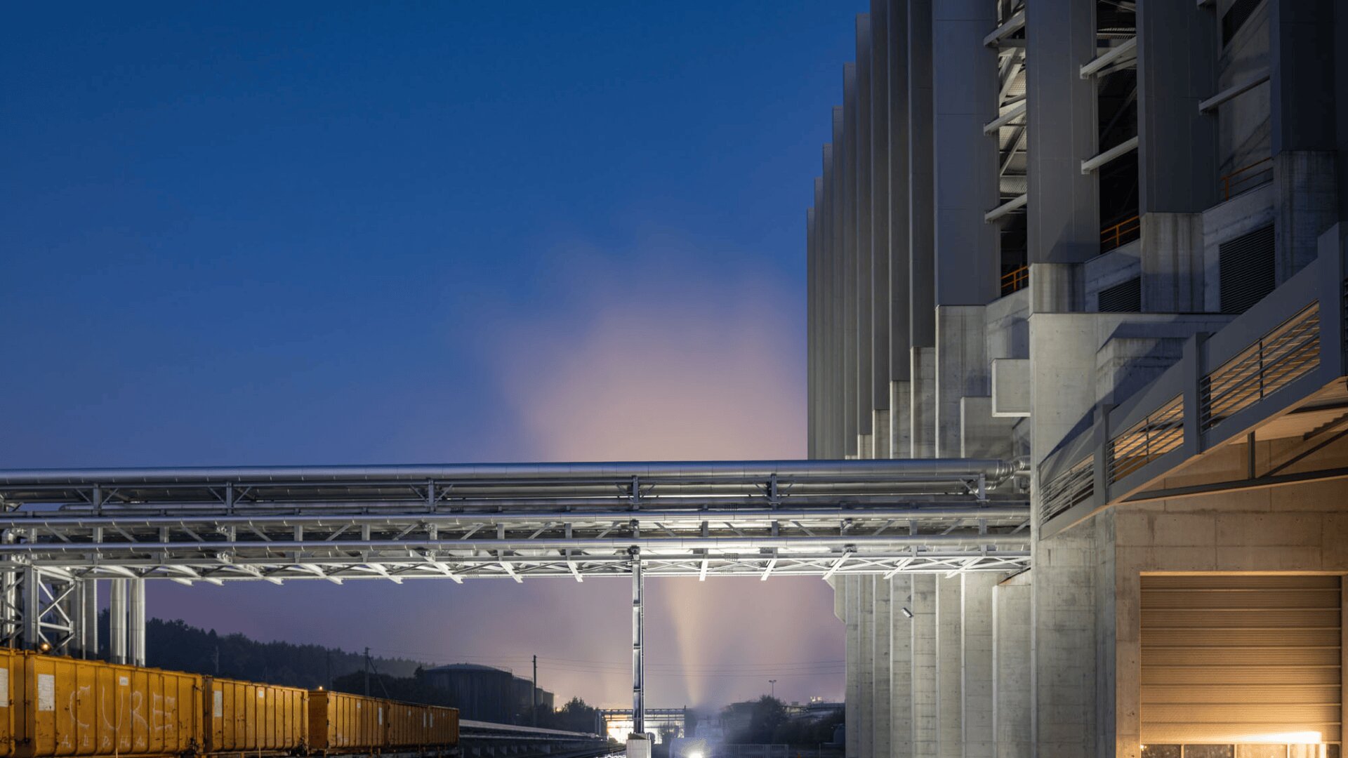 Modern industrial plant at dusk with bright lights illuminating the sky in the background, a metal pipeline running horizontally, yellow train cars below and a large concrete structure on the right side of the image.