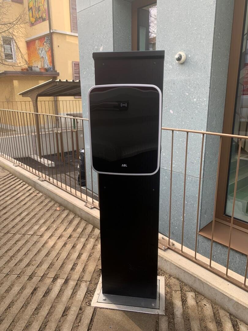 Eine schwarze Ladestation für Elektrofahrzeuge steht auf einem Betonsockel vor einem Gebäude, neben einem Metallgeländer und einem abschüssigen Bürgersteig.