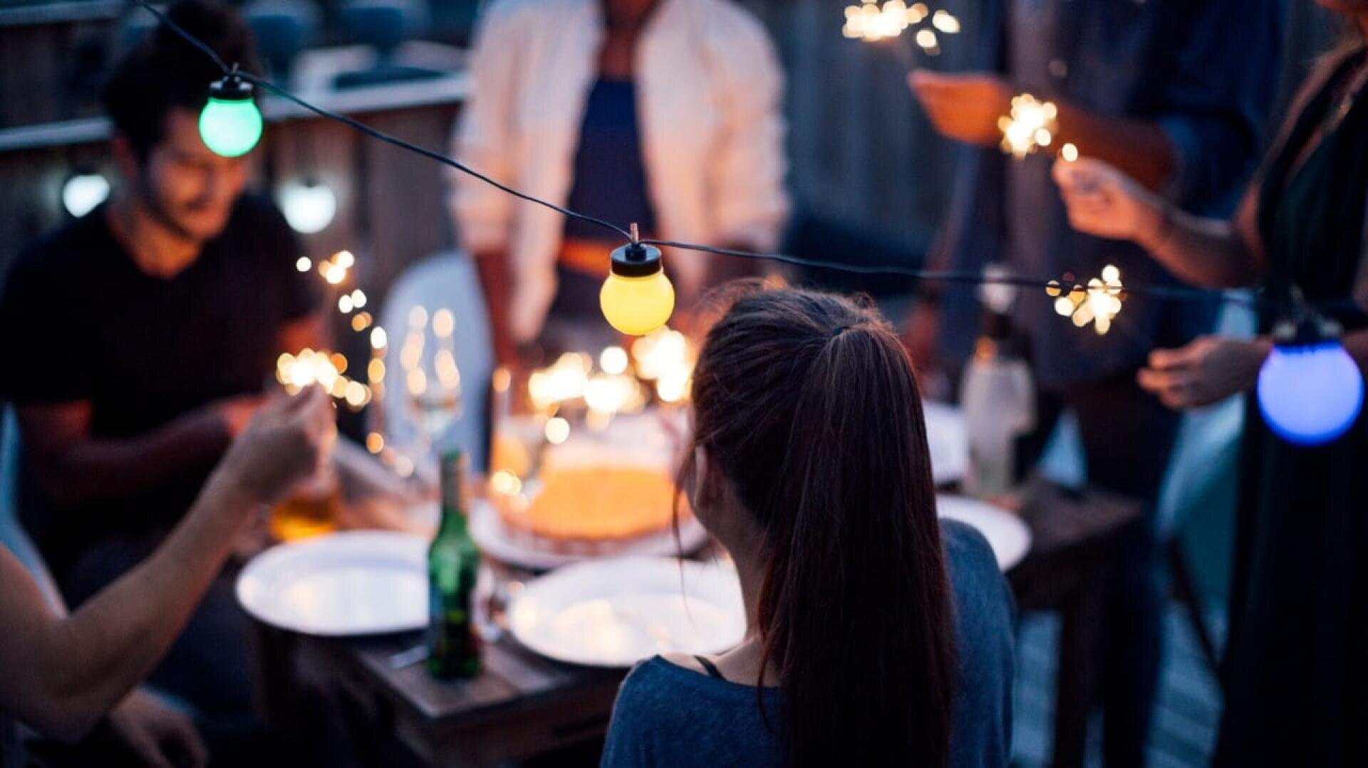 Eine Gruppe von Menschen versammelt sich in der Abenddämmerung um einen Tisch im Freien und hält Wunderkerzen in der Hand. Darüber hängen bunte Lichterketten, und auf dem Tisch stehen ein Kuchen, Teller, Getränke und Flaschen. Die Atmosphäre ist festlich und warm.