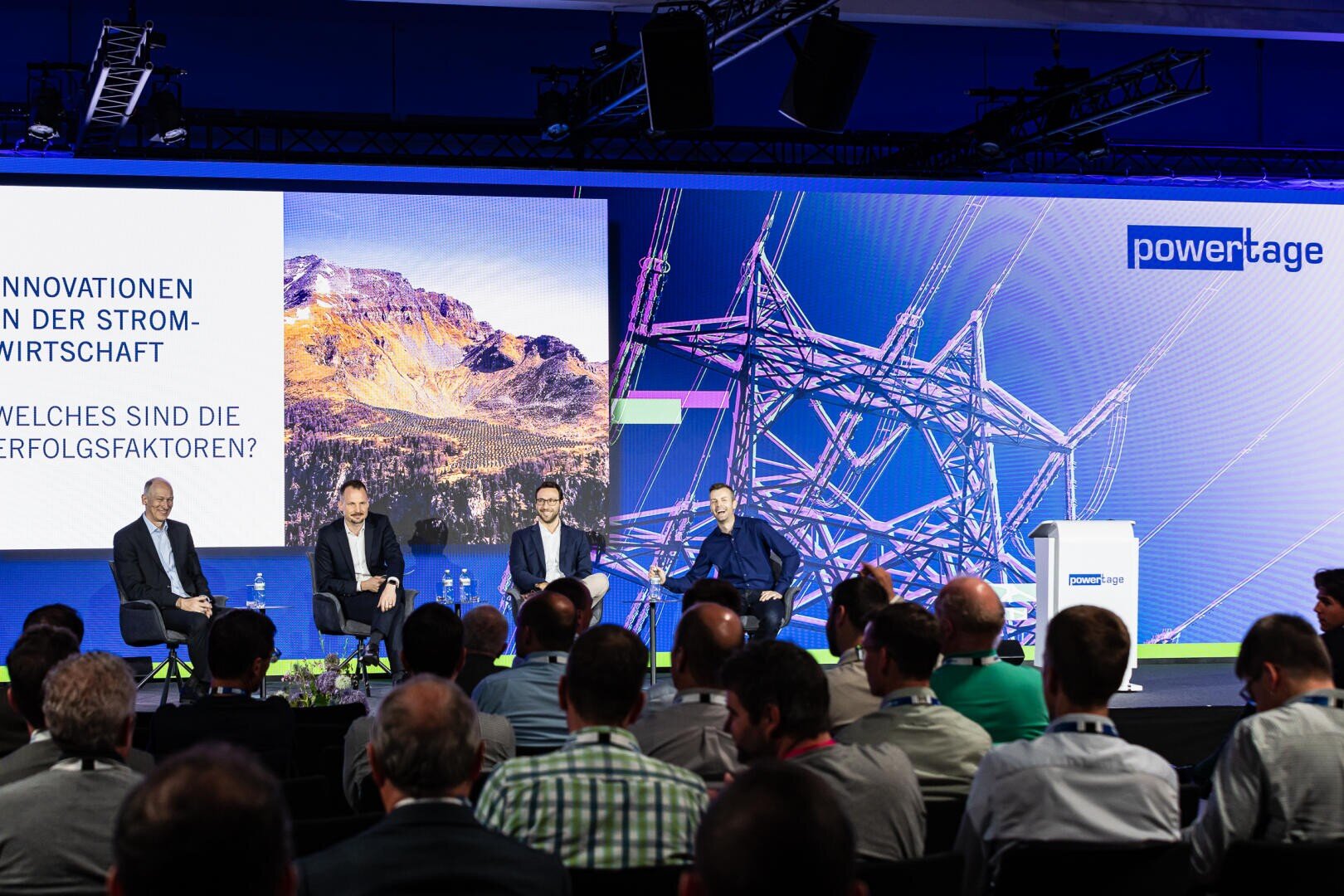 Vier Männer sitzen auf einer Bühne vor einem Publikum bei einer Konferenz. Auf einem großen Bildschirm hinter ihnen sind Berglandschaften, Stromleitungen und ein deutscher Text über Innovationen in der Elektrizitätswirtschaft zu sehen.