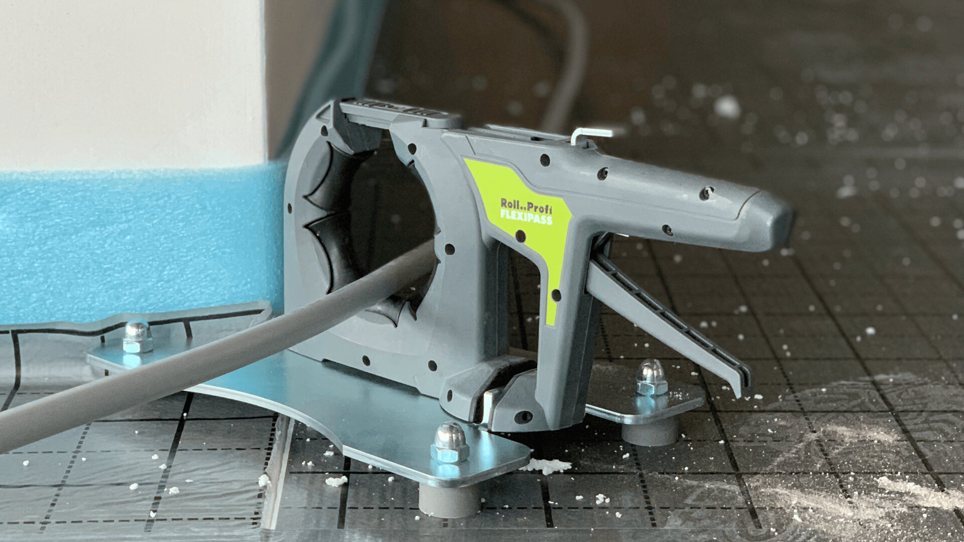 Nahaufnahme eines grauen, handgeführten Kabelhefters mit der Aufschrift Roll-Profi FLEXIPASS, der ein graues Kabel auf einer gitterartigen Oberfläche mit verstreuten weißen Ablagerungen befestigt.