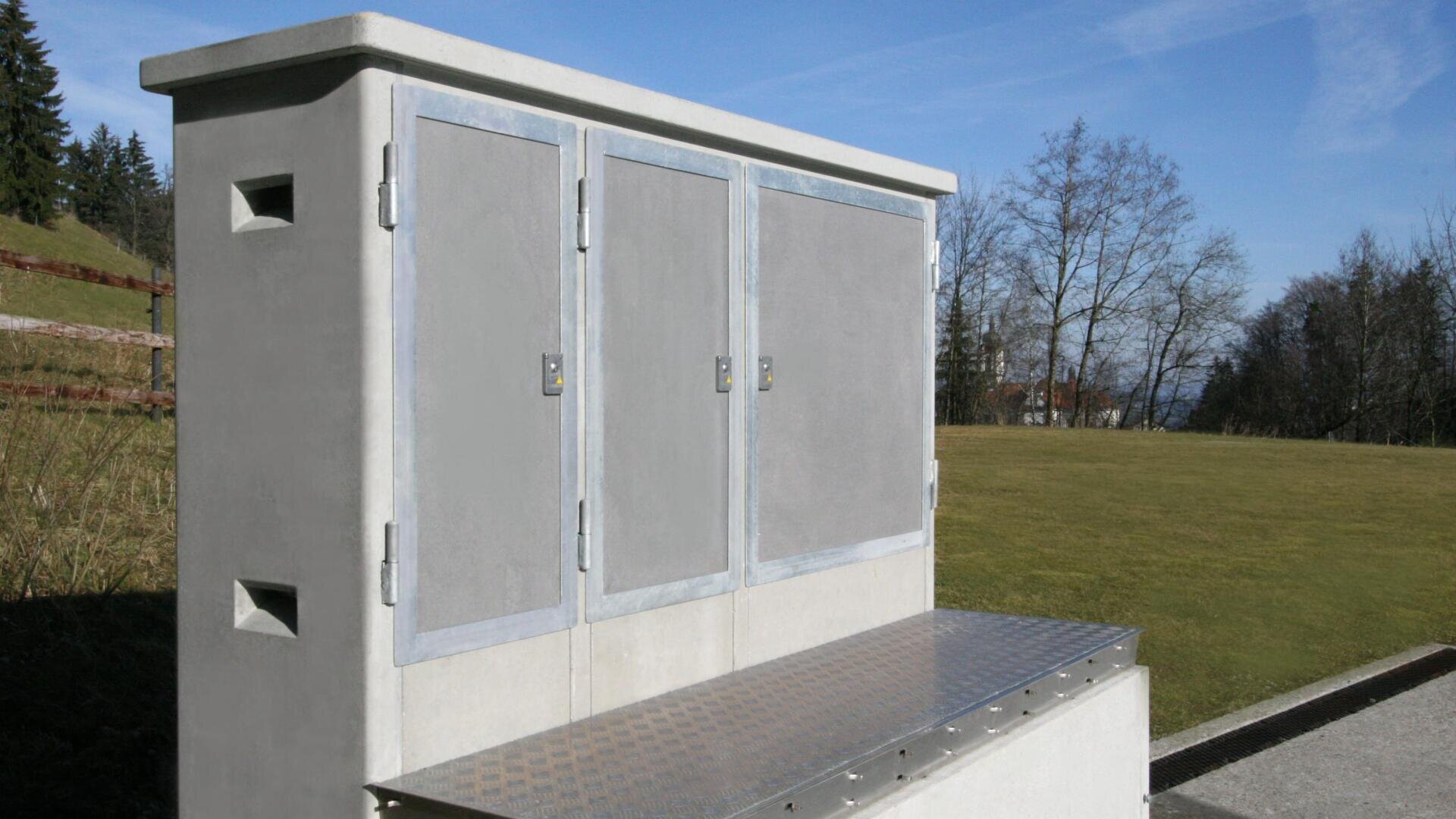 Eine große Außenbox mit drei grauen Metalltüren steht neben einer Wiese und Bäumen unter einem strahlend blauen Himmel.