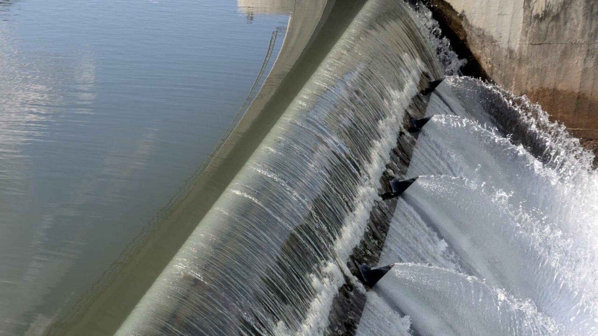 Das Wasser fließt über die Kante eines Betondamms und erzeugt einen sanften, kaskadenartigen Wasserfalleffekt, dessen Spritzer das Sonnenlicht und die umgebende Struktur reflektieren.