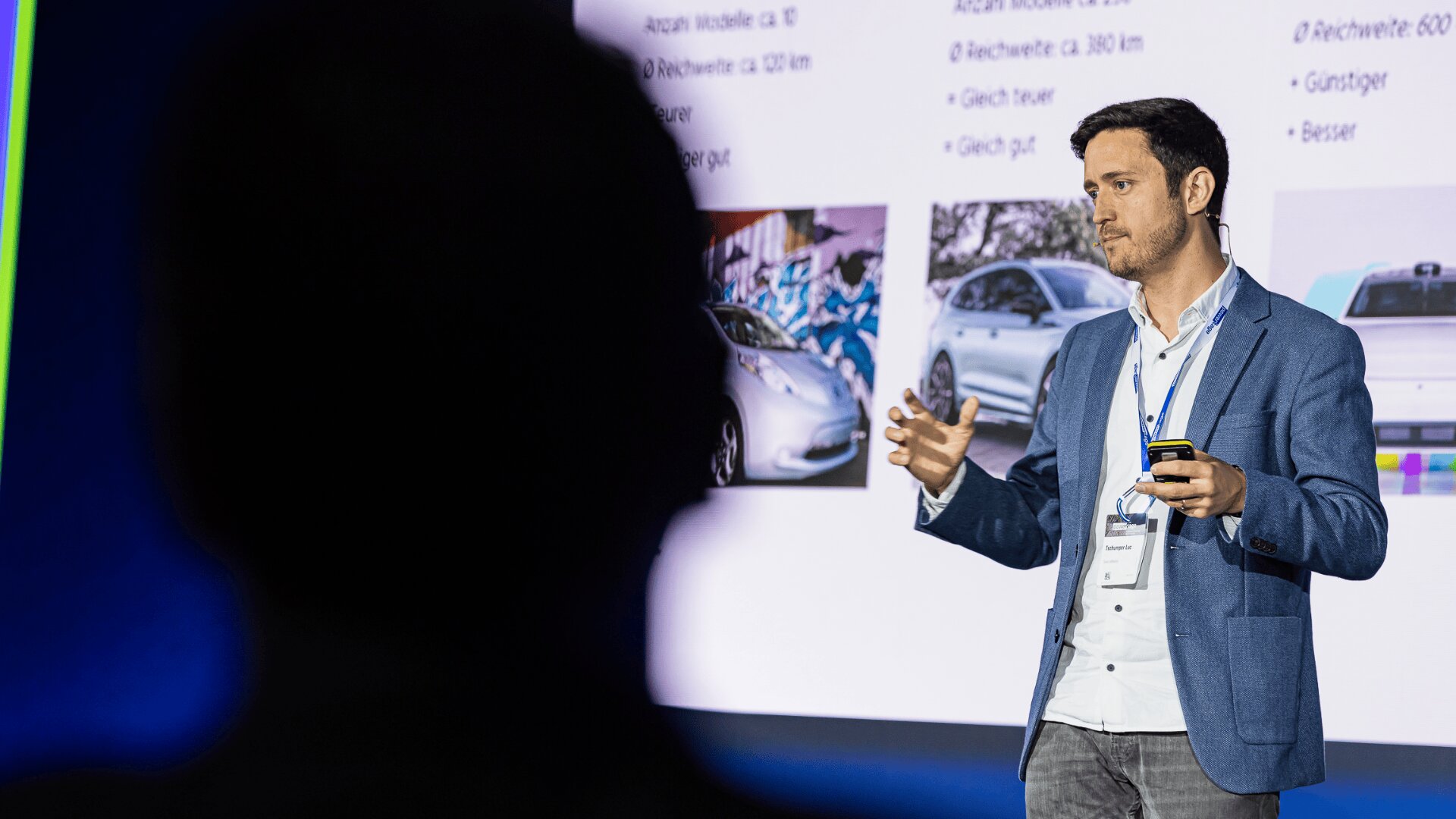 Ein Mann in einem blauen Blazer hält einen Vortrag auf der Bühne und gestikuliert mit seinen Händen. Hinter ihm zeigt ein Bildschirm Bilder von Autos und Text. Im Vordergrund ist die Silhouette eines Zuschauers zu sehen.