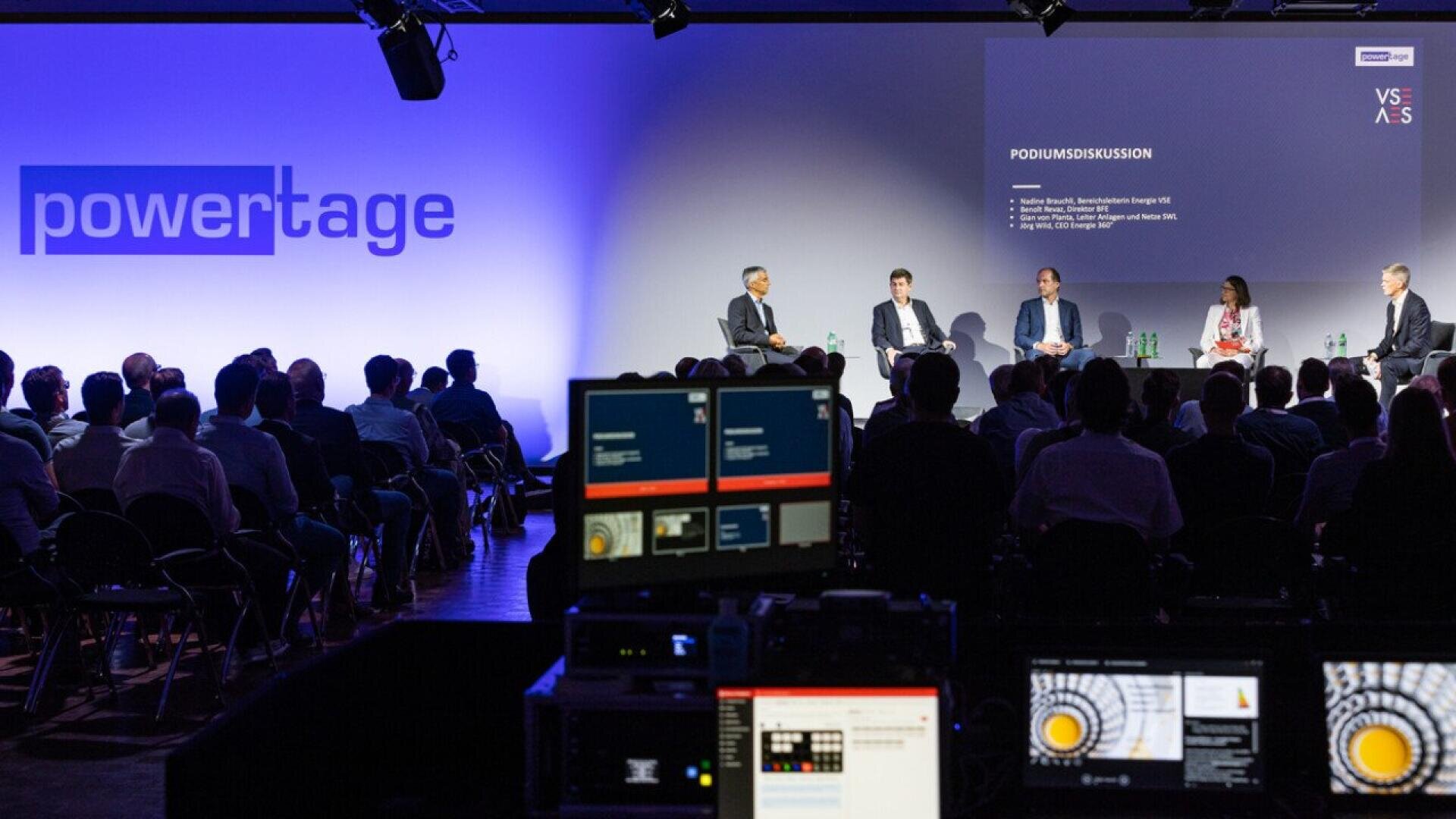 A panel of five people sits on stage in front of an audience at a conference, with a large powertage sign and a podium discussion slide projected on the wall, and audio-visual equipment visible in the foreground.