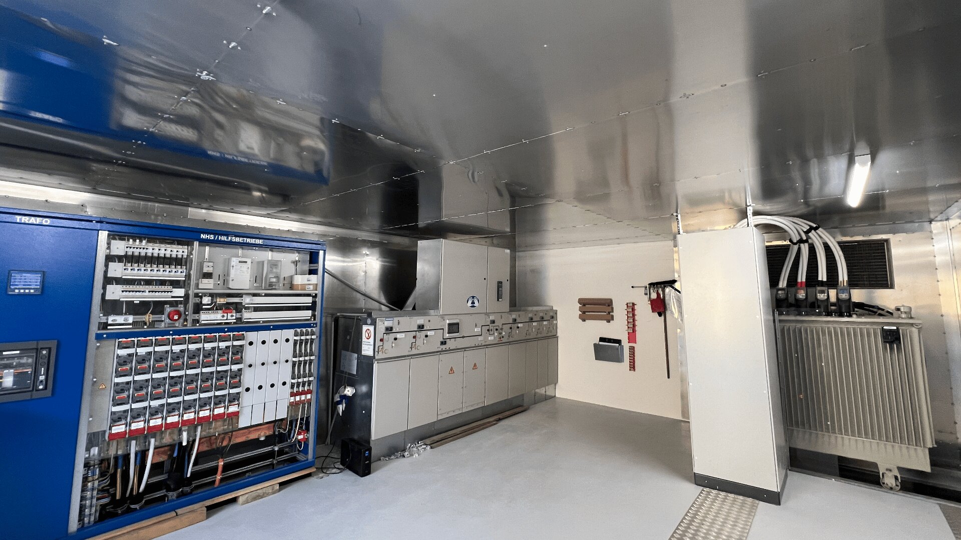 Industrieller Kontrollraum mit einer blauen Schalttafel auf der linken Seite, grauen Schaltschränken in der Mitte und einem großen Transformator auf der rechten Seite. Der Raum hat eine Metalldecke und weiße Wände und Böden.