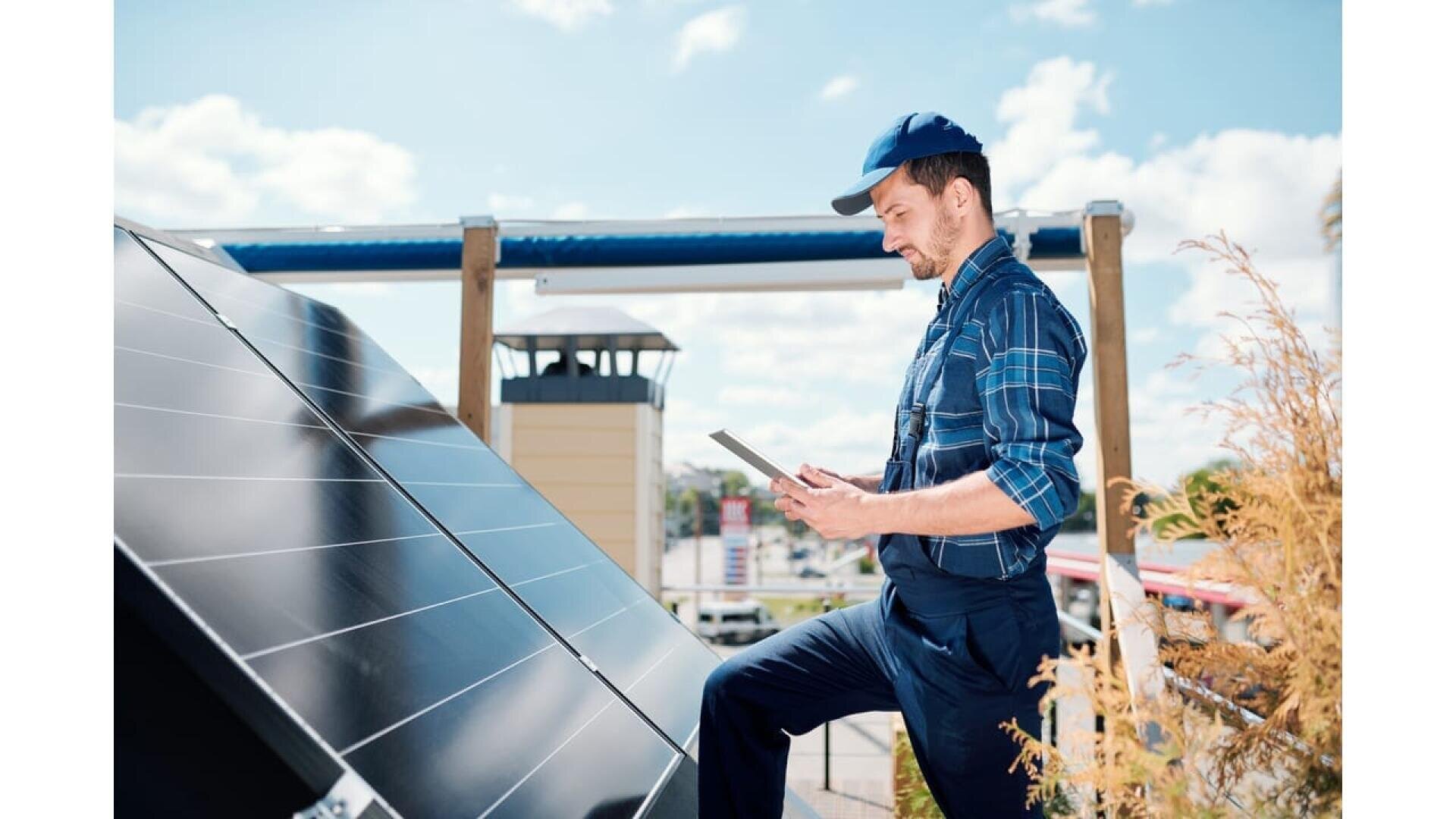 Man in work overalls and cap inspecting solar panels outdoors, holding a digital tablet, with a clear blue sky and buildings in the background.