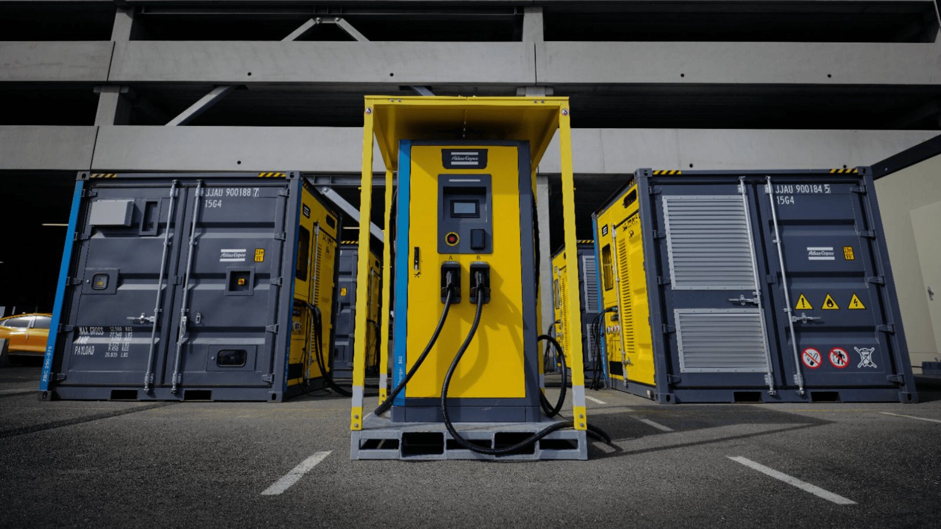 Eine leuchtend gelbe Ladestation für Elektrofahrzeuge mit zwei Ladekabeln steht zwischen zwei großen grauen Industriecontainern auf einem Parkplatz, mit einem Betongebäude im Hintergrund.