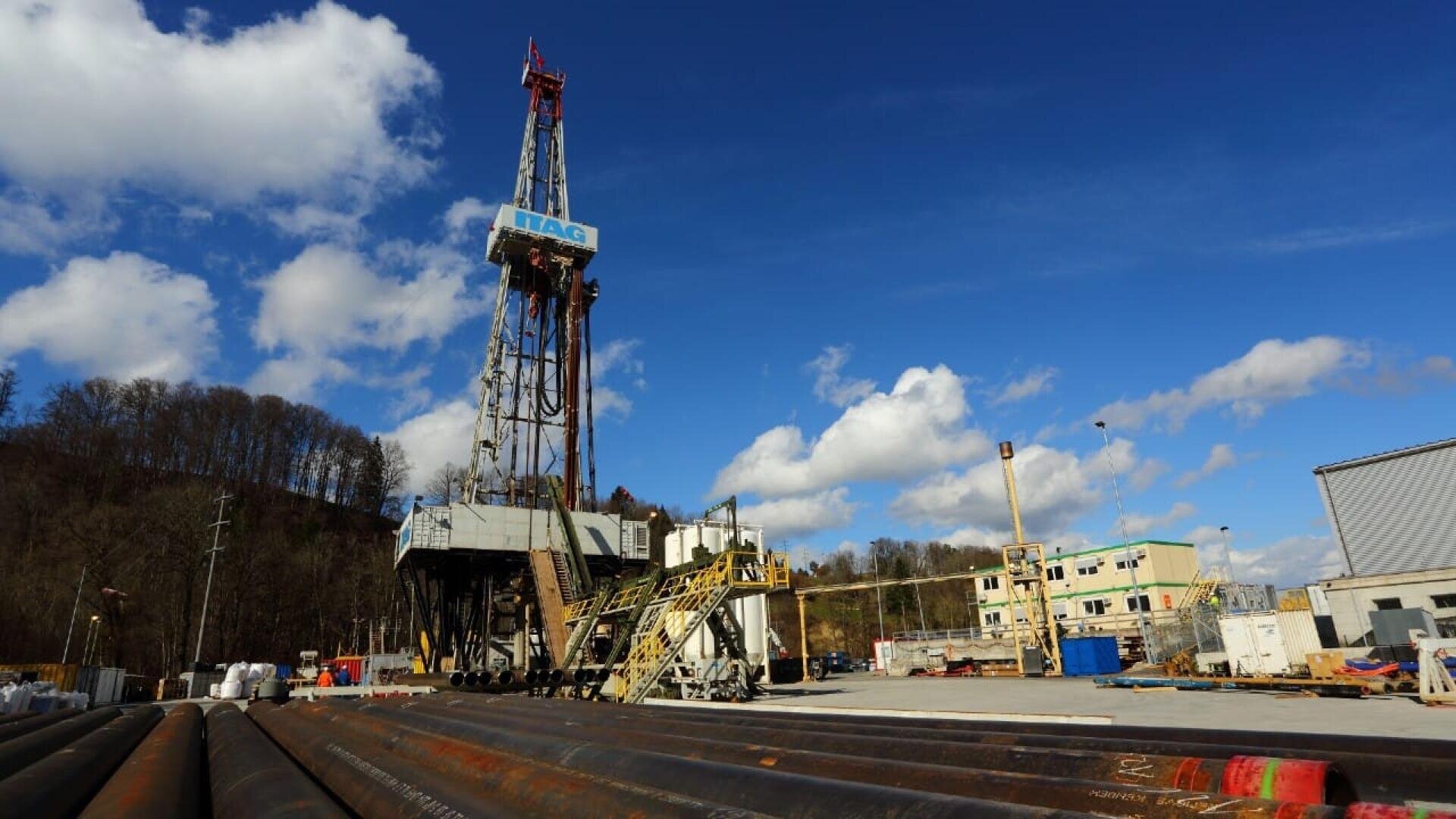 Ein Bohrturm steht hoch auf einem Industriegelände mit Rohren im Vordergrund, Gebäuden und Geräten in der Nähe und einem blauen Himmel mit vereinzelten Wolken über dem Kopf. Bäume säumen einen Hügel im Hintergrund.