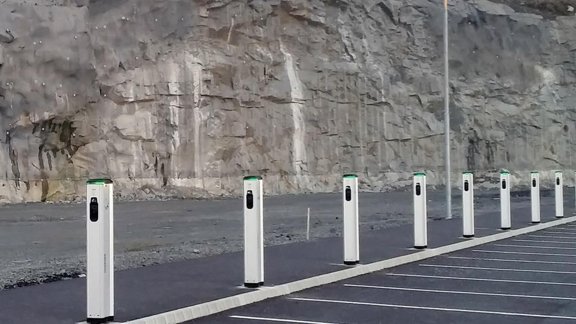 A row of unused electric vehicle charging stations in an empty parking lot, with a rocky cliff face in the background.
