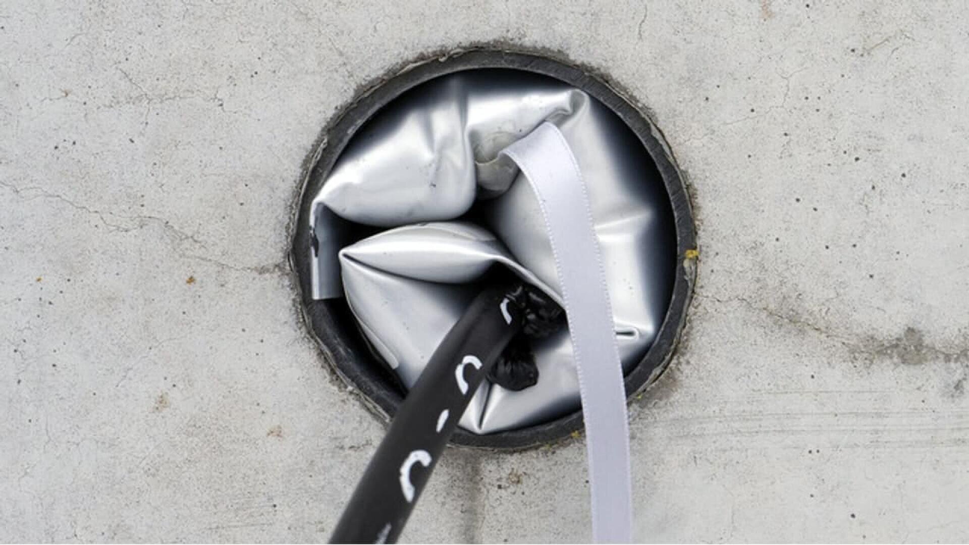 A close-up of a round opening in a concrete surface, partially blocked by a crumpled metallic material. Two cables or ropes—one black and one white—emerge from the opening.