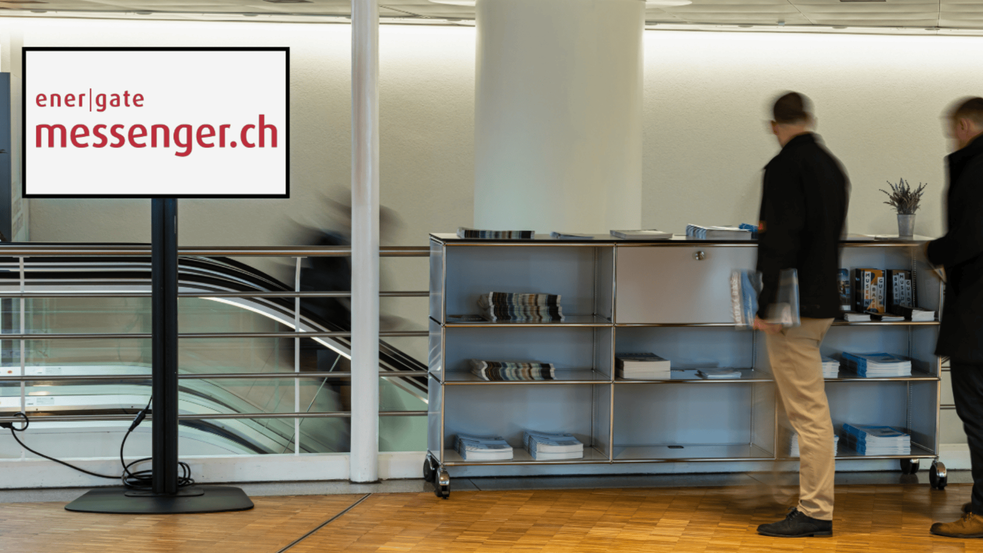 Ein Ständer mit einem Bildschirm mit der Aufschrift "ener|gate messenger.ch" steht neben einem Metallregal mit ordentlich gestapelten Zeitschriften. Zwei unscharfe Personen stehen in der Nähe in einer modernen Inneneinrichtung.