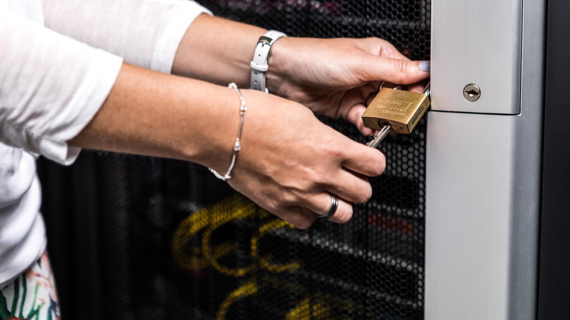 Die Hände einer Person, die einen Serverschrank mit einem Vorhängeschloss verschließt, betonen die Sicherheit und den Schutz von Daten oder Geräten.