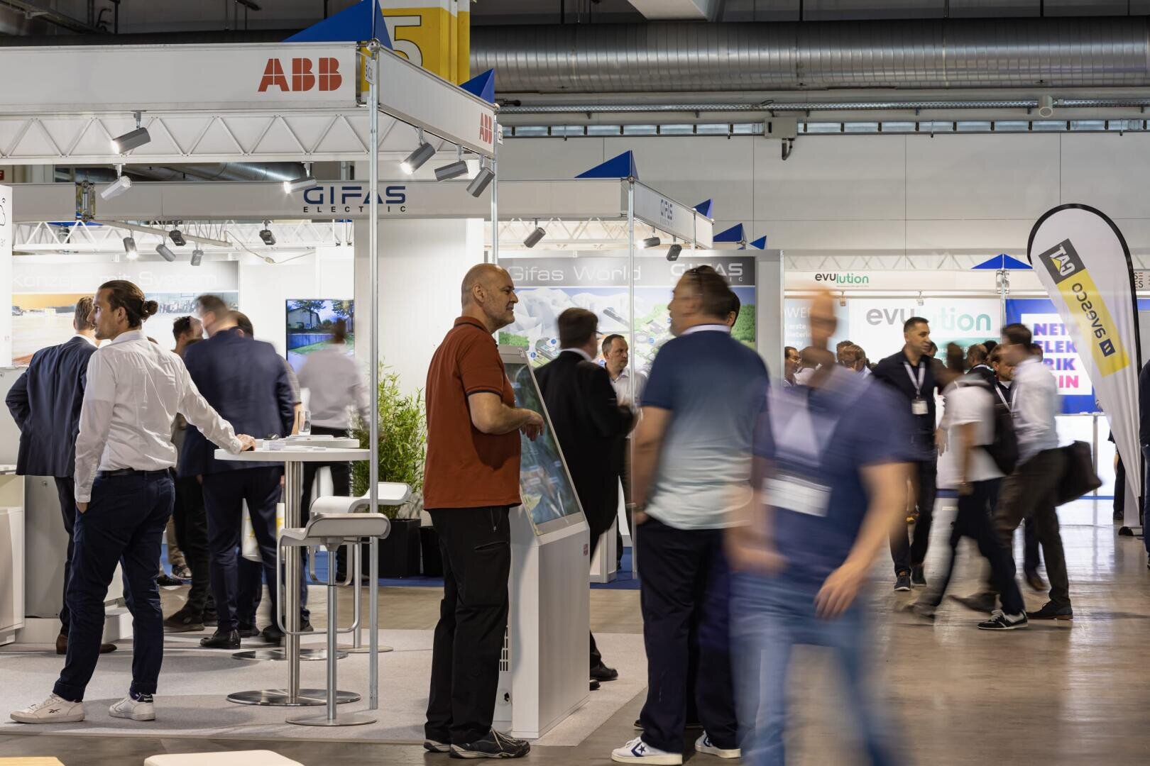 Menschen interagieren und laufen auf einer Messe oder Ausstellung umher, wobei im Hintergrund verschiedene Stände, Banner und Displays zu sehen sind. Einige Menschen sind in ein Gespräch vertieft, während andere sich durch den Raum bewegen.