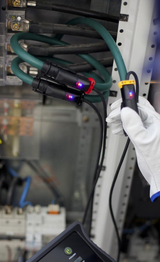 A person wearing a white glove holds a cable with a sensor attached to thick green and black electrical wires inside an electrical panel. Small red and blue lights are visible on the connectors.
