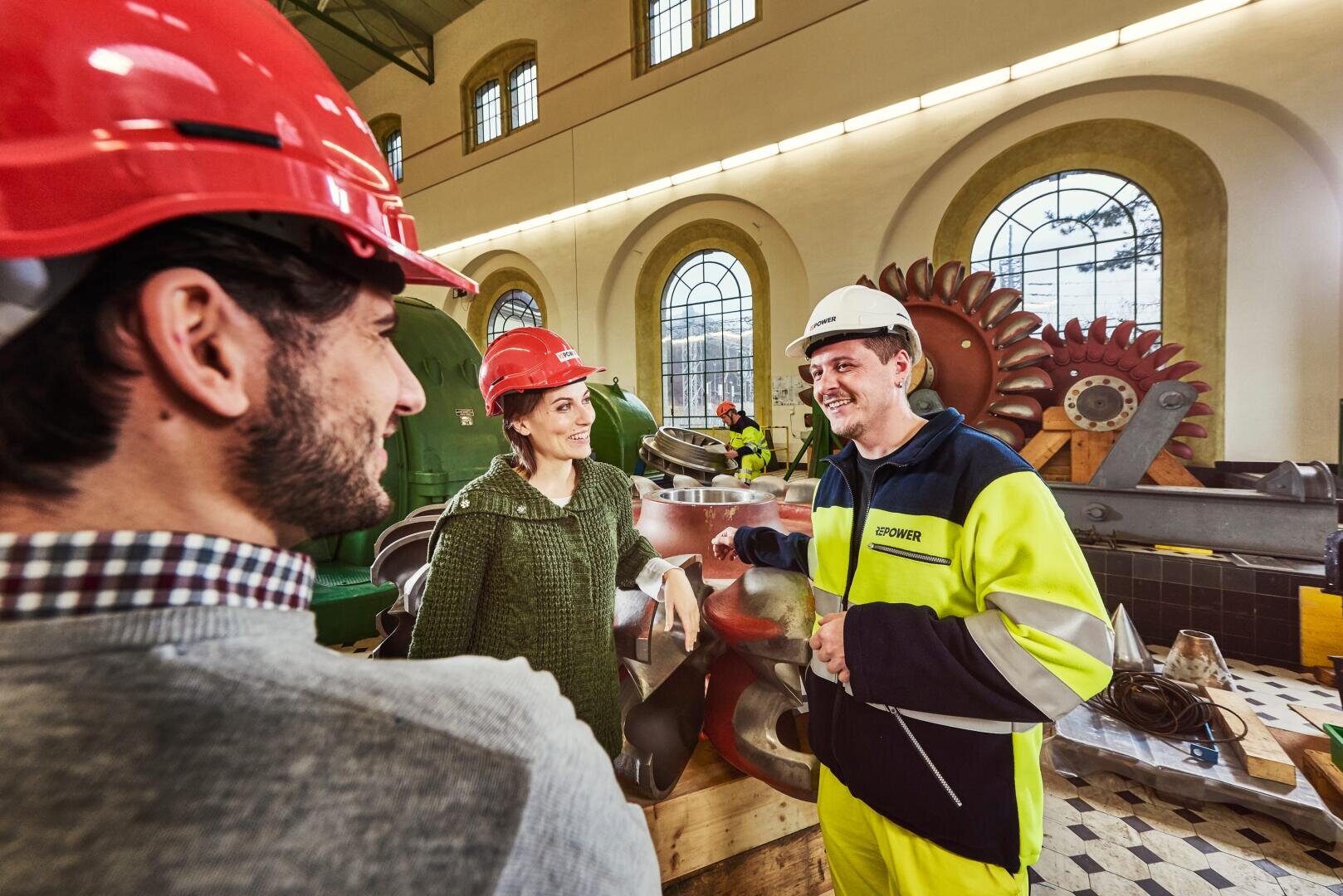 Drei Personen mit Schutzhelmen und Sicherheitsausrüstung stehen in einem Innenraum neben Industriemaschinen, lächeln und unterhalten sich. Große gewölbte Fenster lassen Tageslicht herein, und im Hintergrund sind Turbinenteile zu sehen.