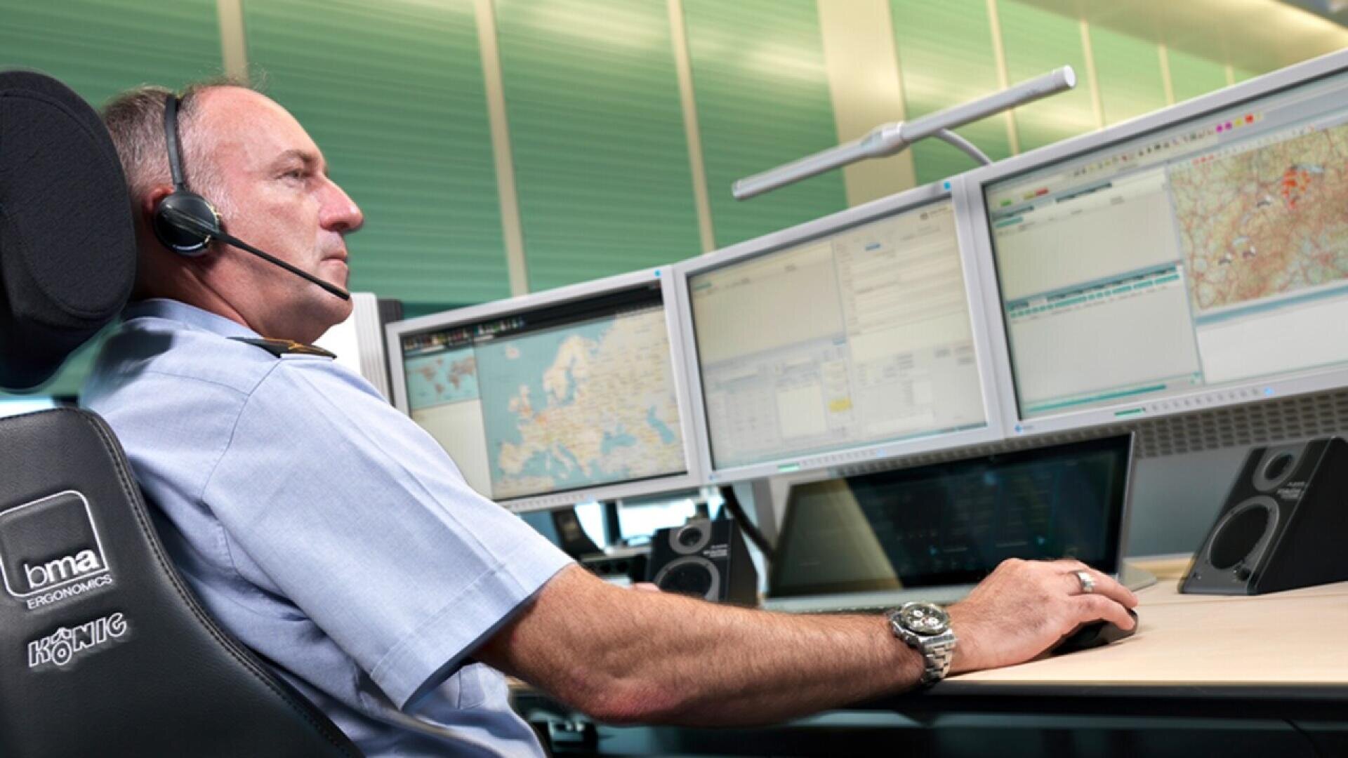 A man wearing a headset sits at a workstation with four computer monitors displaying maps and data, appearing focused as he manages information in a modern control room setting.