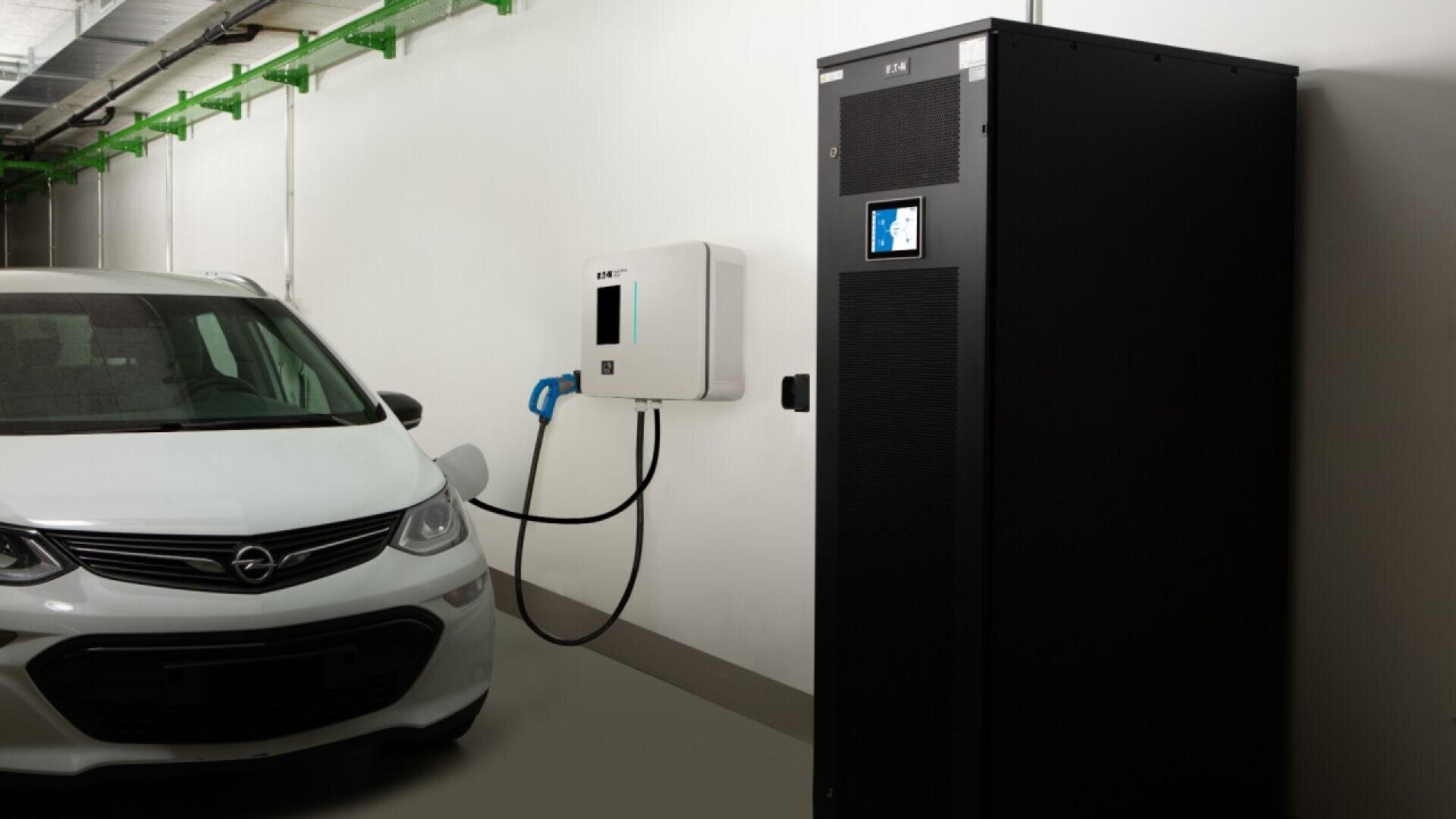 A white electric car is parked indoors next to a wall-mounted EV charging station, which is connected to the car. A tall black cabinet with a digital display stands nearby.