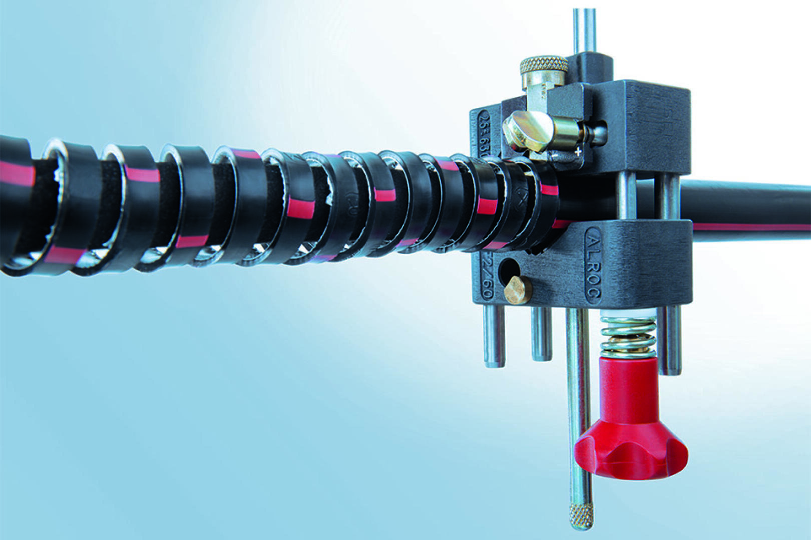 Close-up of a mechanical device with a black and red spiral-wrapped cable, metal rods, adjustment knobs, and a red component against a blue background. The setup appears technical or industrial.