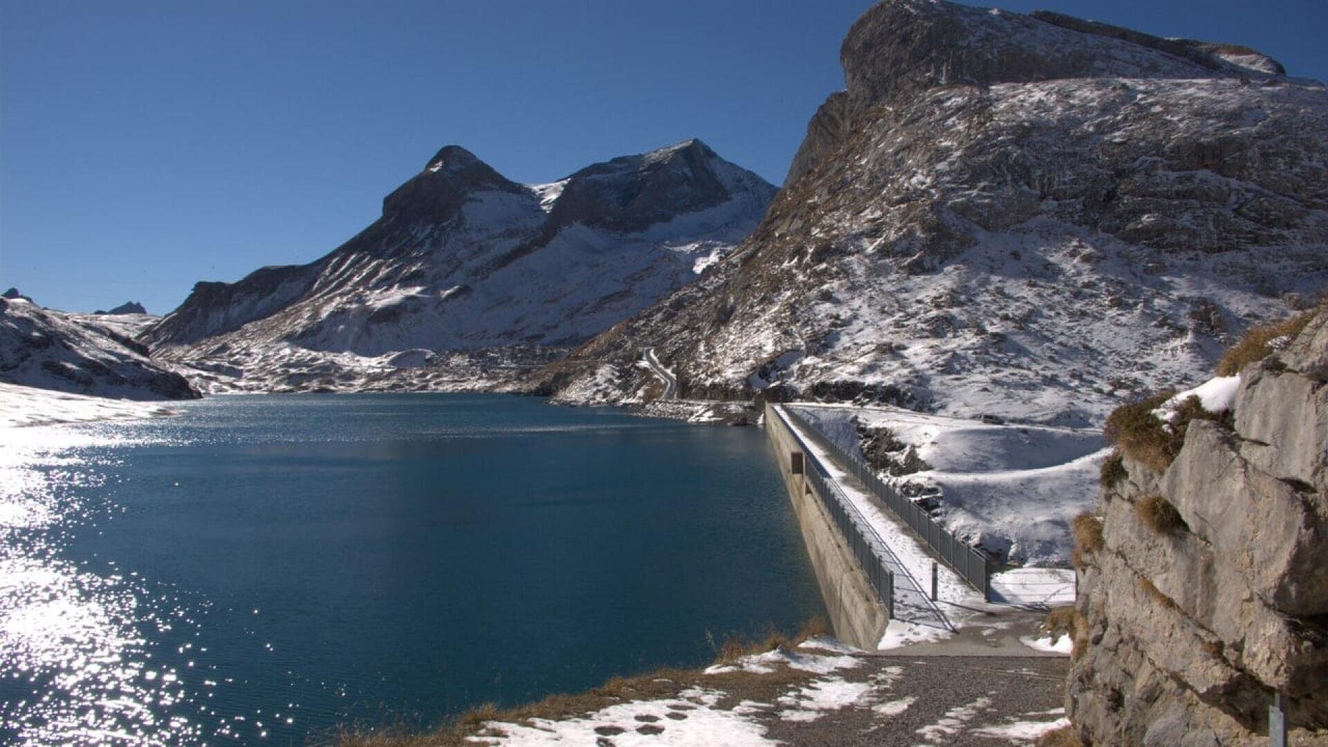 A clear blue reservoir is bordered by a concrete dam, surrounded by snow-dusted rocky mountains under a bright blue sky. Sunlight reflects off the water, creating a serene and crisp winter scene.