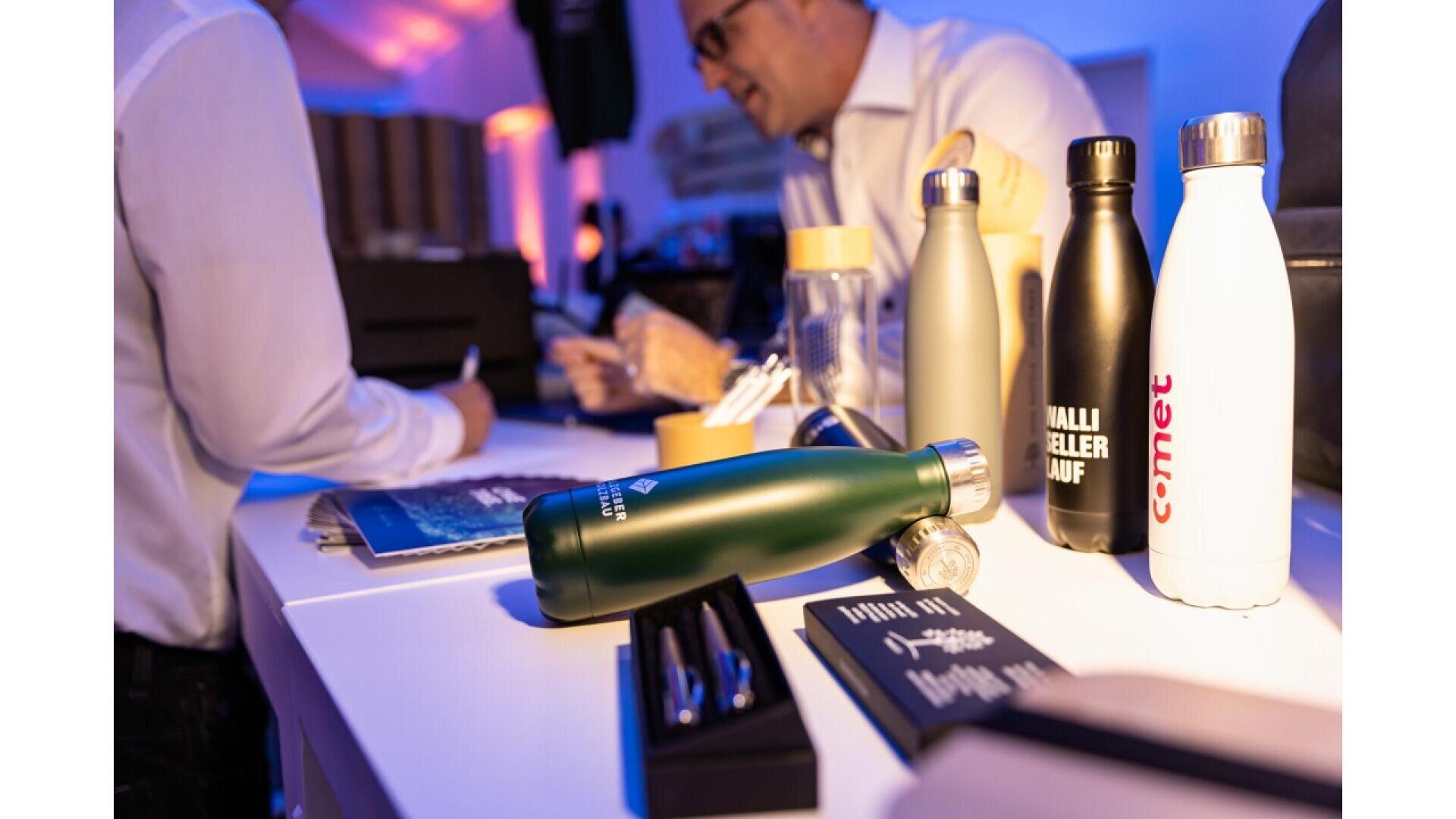 A close-up of reusable water bottles and pens displayed on a table, with two people in the background interacting in a well-lit indoor setting.