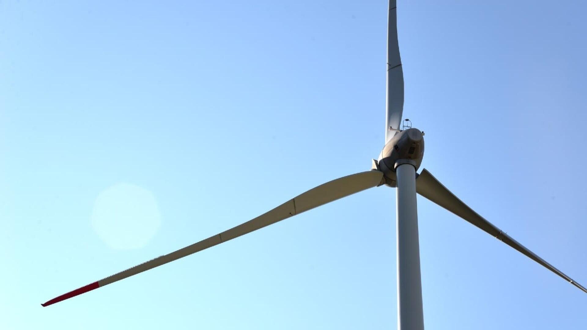 Nahaufnahme einer Windkraftanlage mit drei Flügeln, von denen einer rot ist, vor einem klaren blauen Himmel an einem sonnigen Tag.