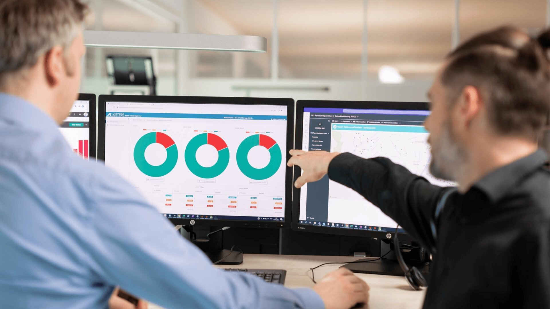 Zwei Personen in einem Büro schauen auf Computermonitore, auf denen kreisförmige Diagramme und Datentabellen angezeigt werden. Eine Person zeigt auf den Bildschirm, während sie über die angezeigten Informationen spricht.