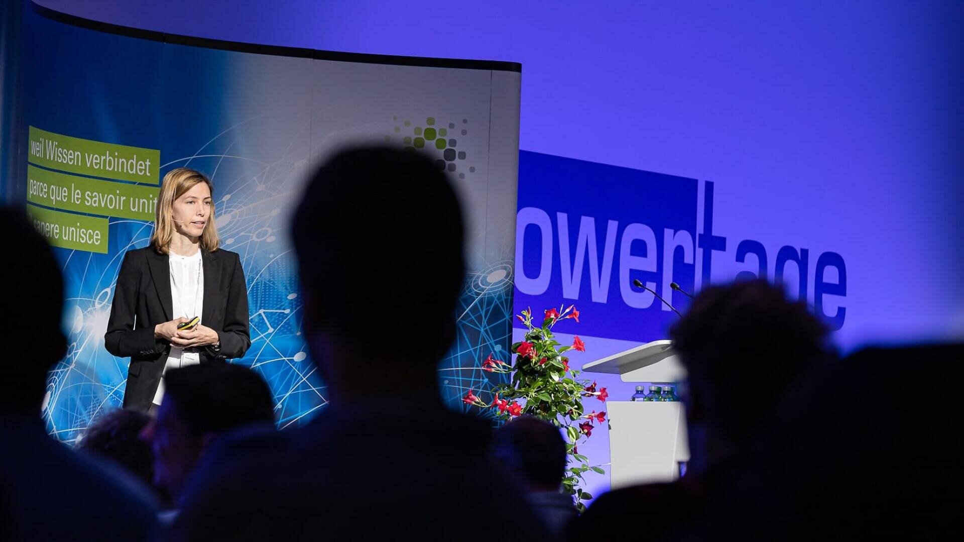 Eine Frau in einem Blazer spricht auf der Bühne einer Konferenz mit blauem Hintergrund, Präsentationsbildschirm und Podium mit Blumen. Die Zuhörer sind im Vordergrund zu sehen.