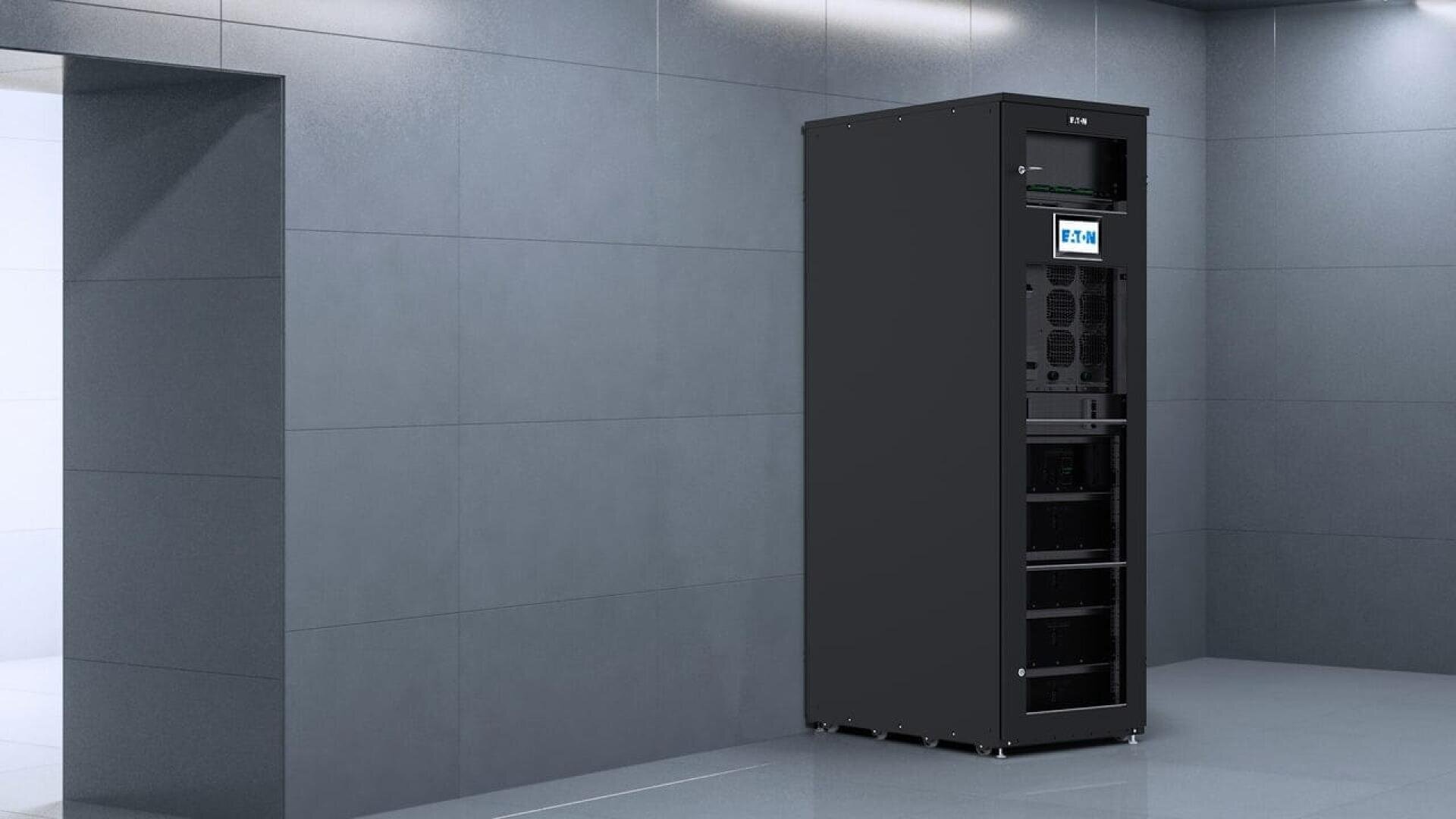 A tall, black server rack with electronic components and a digital display stands against a gray tiled wall in a modern, empty room.