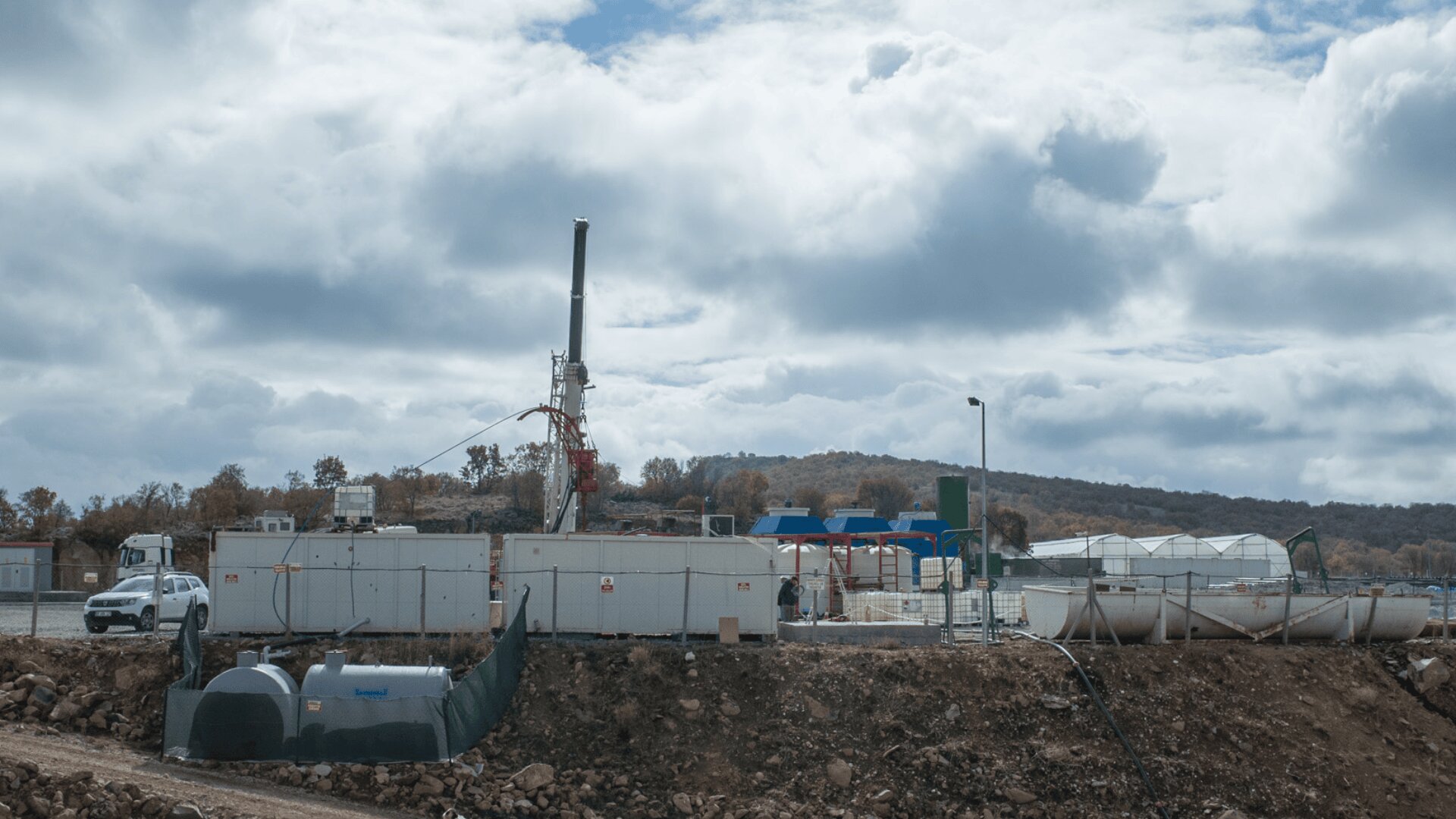 A natural gas drilling site with a tall rig, several white container units, storage tanks, and vehicles under a cloudy sky, surrounded by a fence and rocky ground. Trees and hills are visible in the background.