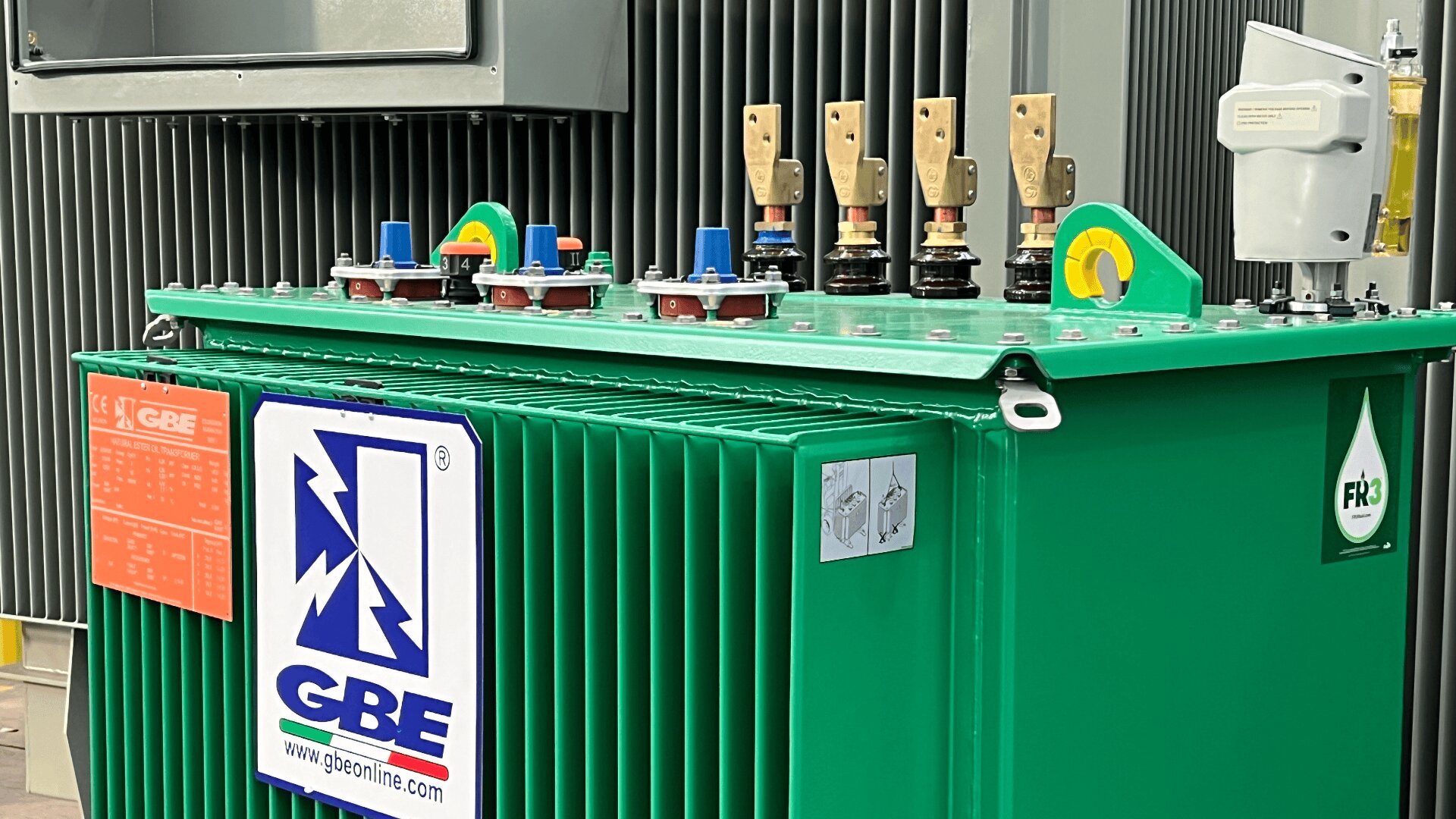 A green GBE electrical transformer with various colored terminals and switches on top, labeled with a blue and white GBE logo. Large vertical metal fins and an orange information plate are visible in the background.