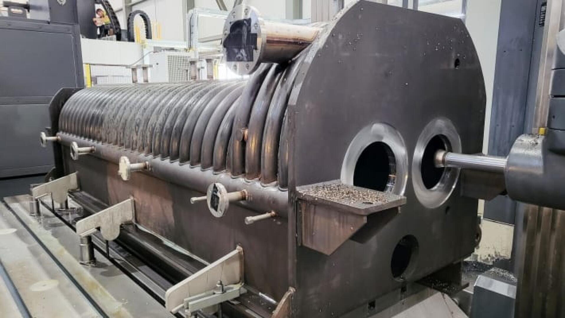 Eine große, zylindrische Industriemaschine aus Metall wird bearbeitet, wobei an einer offenen Öffnung Metallspäne zu sehen sind. Die Maschine hat eine gerippte Oberfläche und befindet sich in einer Fabrikumgebung mit verschiedenen Geräten im Hintergrund.