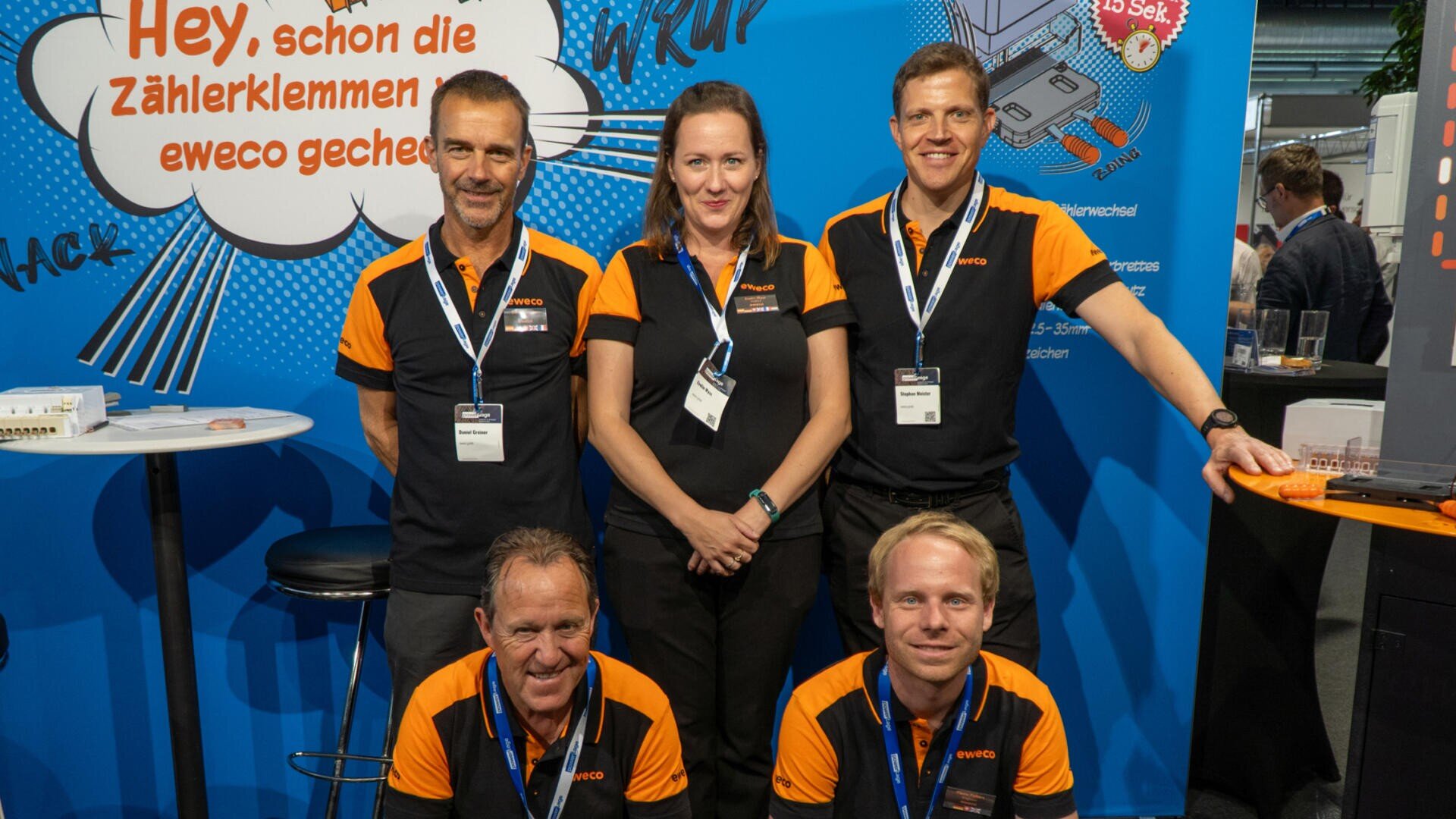 Five people in black and orange uniforms pose and smile in front of an exhibition stand with a light blue background, on which German texts and illustrations on the subject of technology can be seen.
