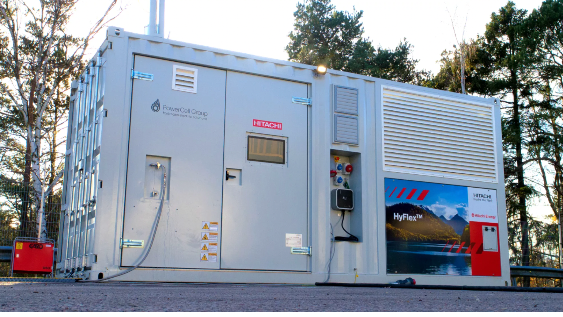 Ein großer, weißer Industriecontainer mit der Aufschrift PowerCell Group, Hitachi und HyFlex, mit Kabeln und Schalttafeln, steht unter freiem Himmel zwischen Bäumen.