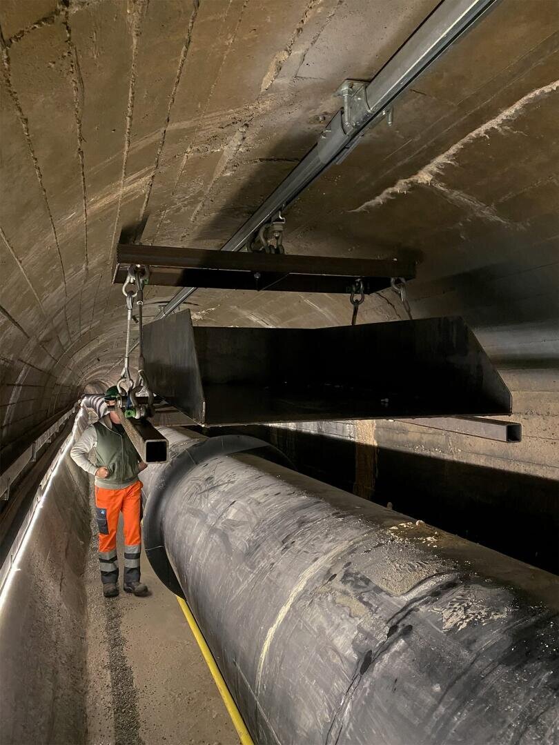 Ein Arbeiter in Schutzausrüstung steht in einem Betontunnel neben einem großen Rohr und beobachtet eine aufgehängte Metallplattform, die mit Ketten und Flaschenzügen an der Tunneldecke befestigt ist.