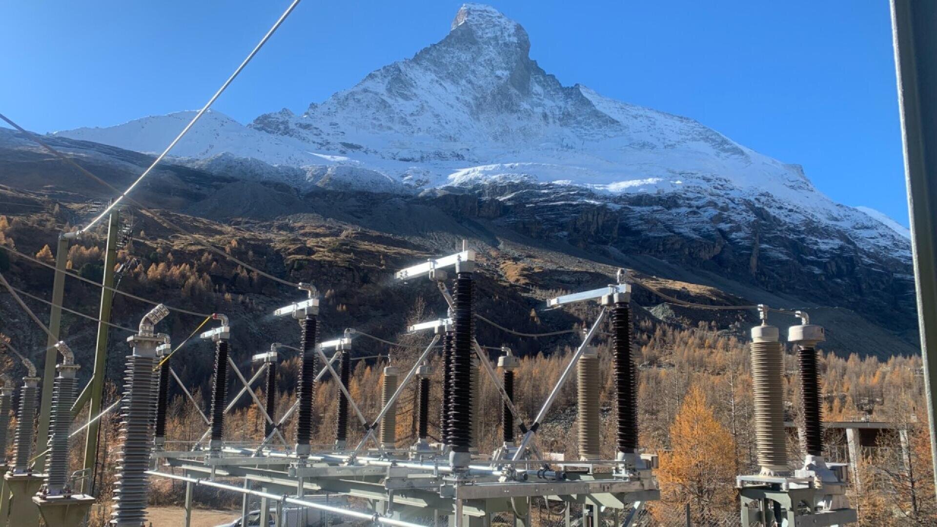 An electrical substation with tall insulators stands in the foreground, set against a backdrop of snow-capped mountains and autumn trees under a clear blue sky.
