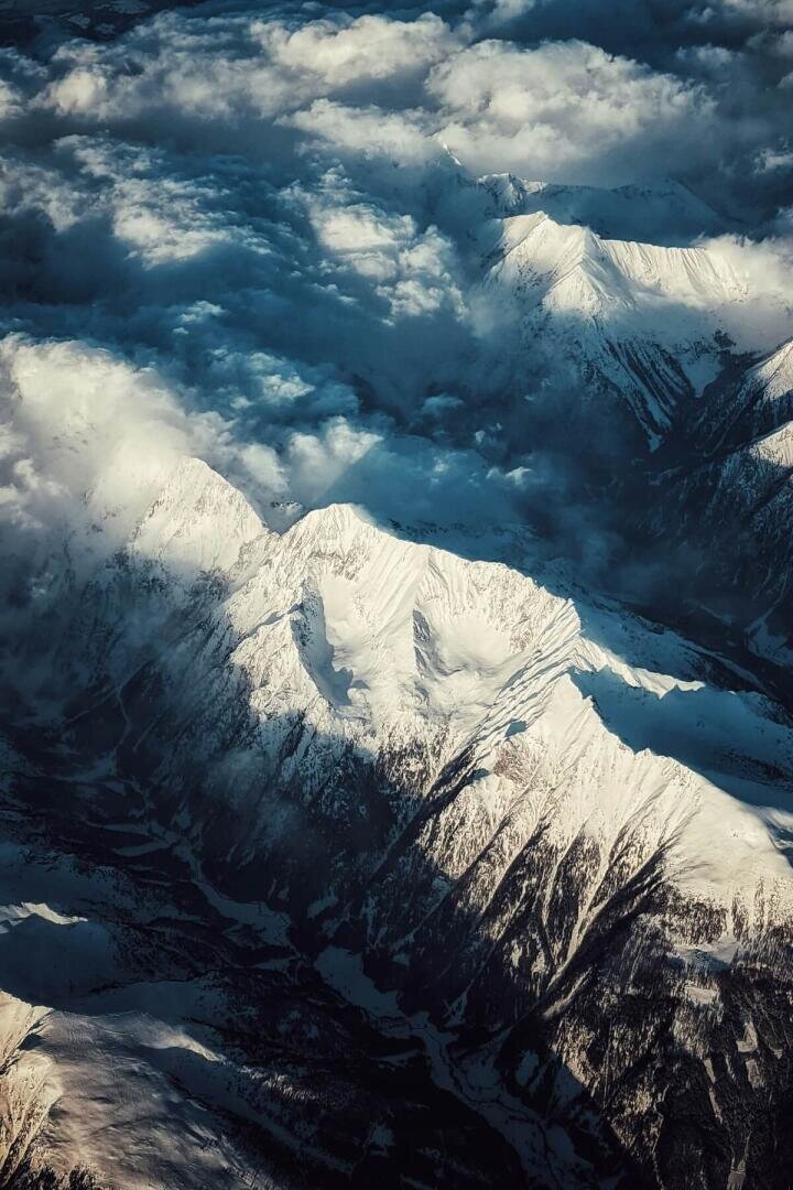Luftaufnahme von schneebedeckten Berggipfeln, die teilweise von Wolken verhüllt sind, während das Sonnenlicht dramatische Schatten auf die zerklüftete Landschaft wirft.