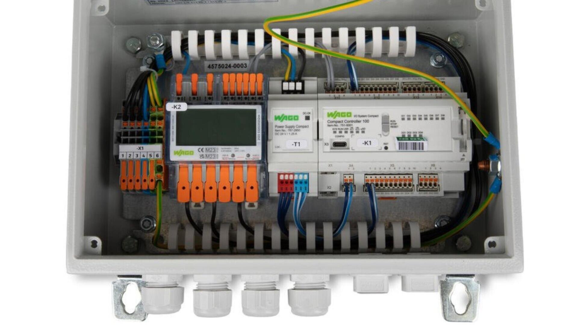 An open electrical control box showing various wires, connectors, and electronic modules mounted on a DIN rail, with labeled terminals and organized wiring inside a grey enclosure.