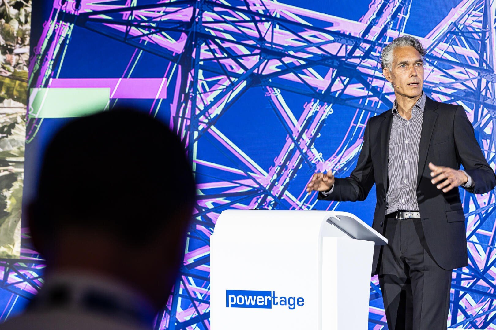 Ein Mann im Anzug spricht auf einem Podium mit der Aufschrift "Powertage" vor einem farbenfrohen, abstrakten Hintergrund mit Stromleitungsstrukturen hinter ihm. Eine weitere Person ist im Vordergrund als Silhouette zu sehen.