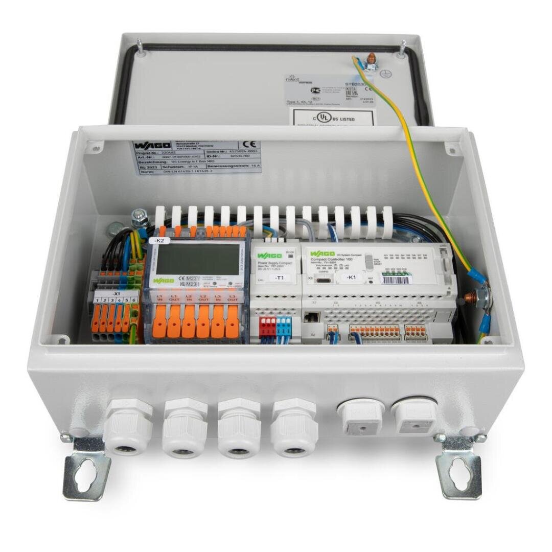 Ein weißer Schalttafelkasten mit geöffnetem Deckel, in dem verschiedene Kabel, Steckverbinder, orange und grün beschriftete Klemmenleisten und elektronische Bauteile übersichtlich angeordnet sind. An der Unterseite sind vier Kabelverschraubungen zu sehen.