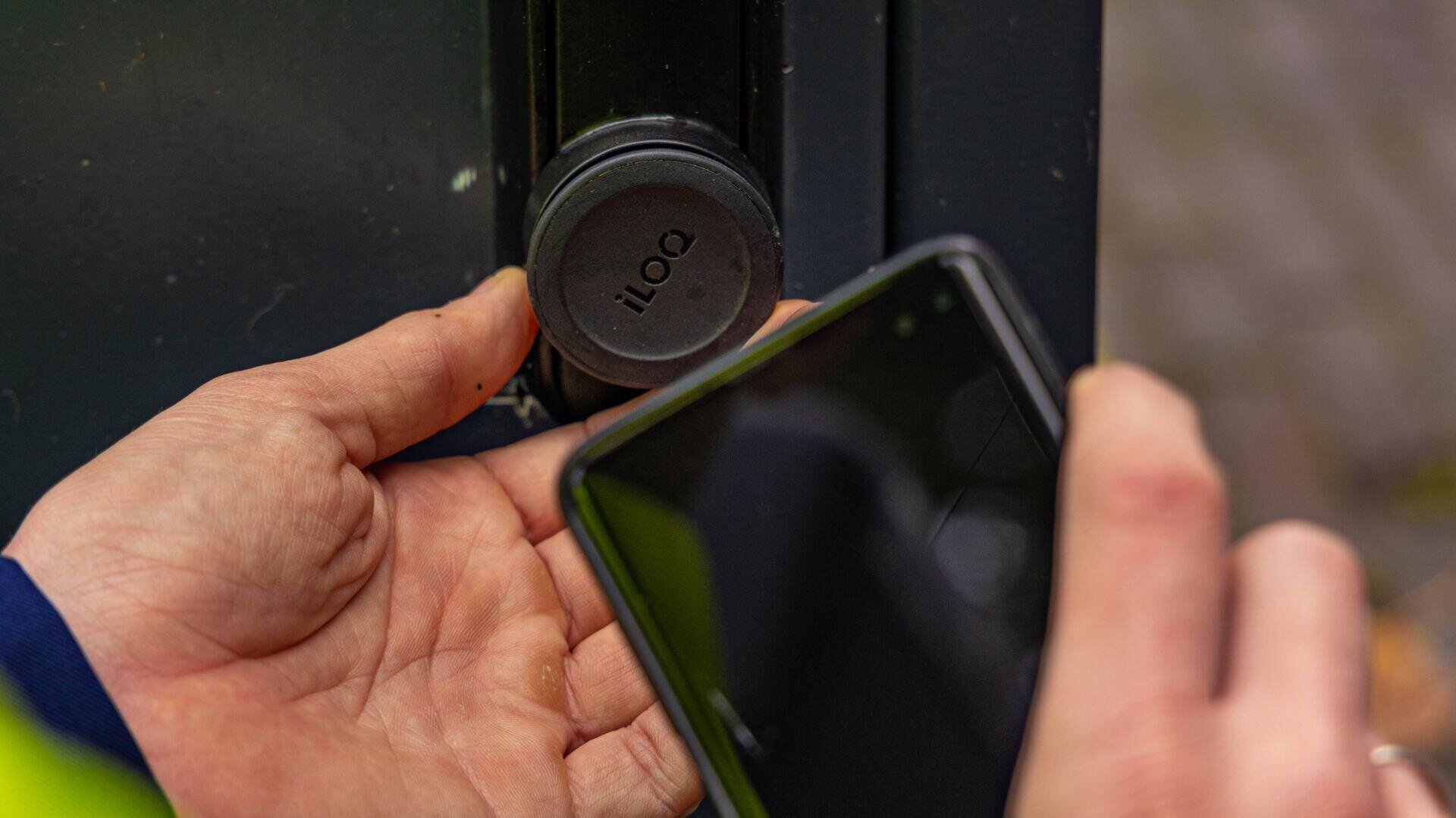 A person holds a smartphone near an iLOQ smart lock, preparing to unlock a door using digital access. The persons hand is close to the lock, and the phone screen is visible.