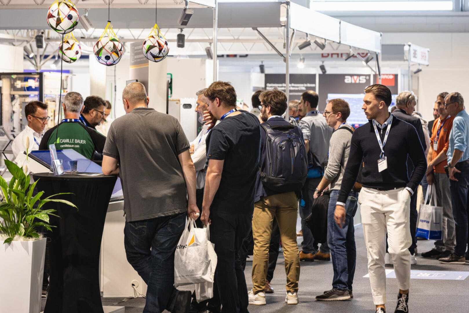 Eine Gruppe von Menschen steht in einer Schlange an einem belebten Messestand. Einige halten Tragetaschen oder tragen Anstecker, während über dem Stand Fußbälle hängen. Es gibt Auslagen, eine Pflanze und helle Innenbeleuchtung.
